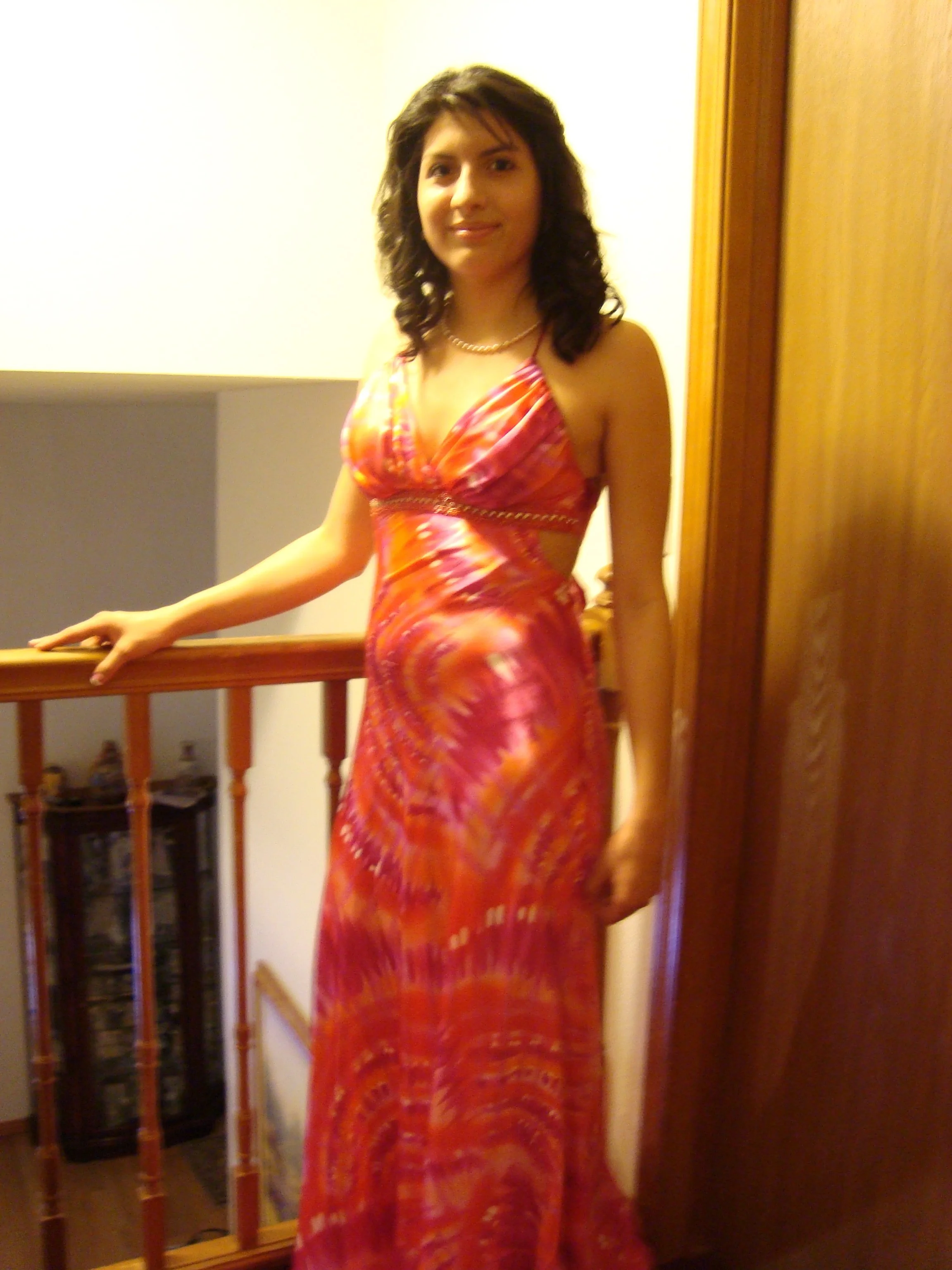 A woman with dark hair in a colorful, patterned dress standing on a staircase with wooden railings inside a house.