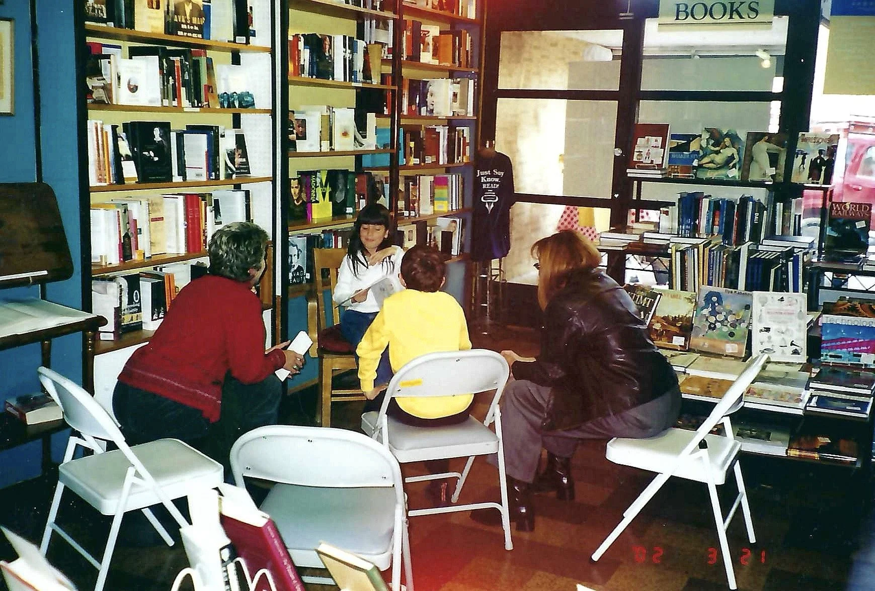A group of people sitting around a table inside a bookstore, engaging in a discussion or book reading activity. Bookshelves filled with books line the walls, and a sign that says "BOOKS" is visible in the upper right corner.