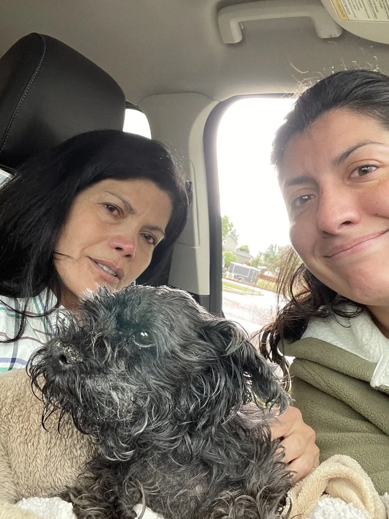 A woman, a young girl, and a black curly-haired dog sitting in a vehicle, with the woman and girl smiling at the camera.
