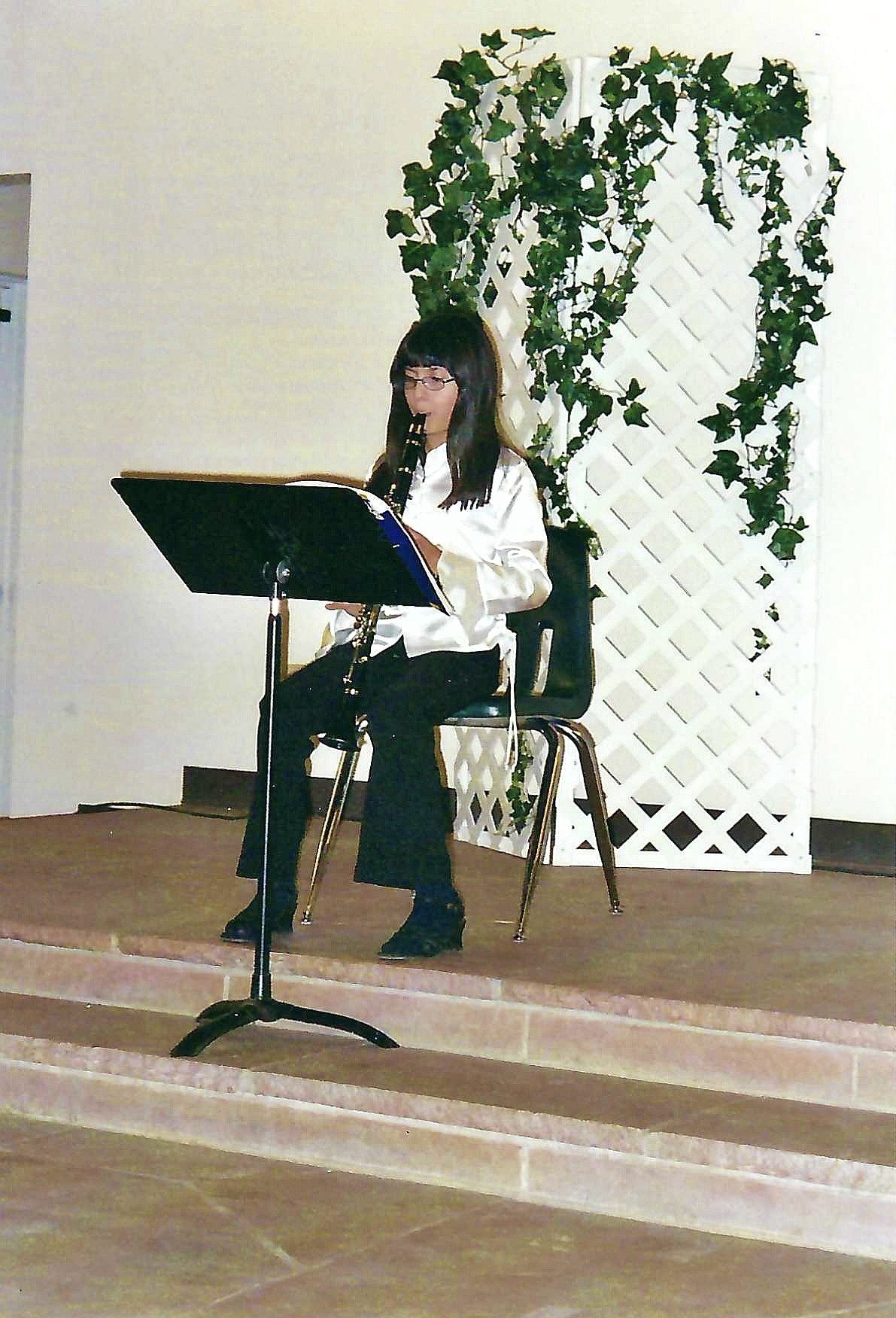 A young Asian woman with glasses playing the clarinet on a small stage, seated on a chair with a music stand in front of her, and a decorative white lattice with green ivy plants behind her.