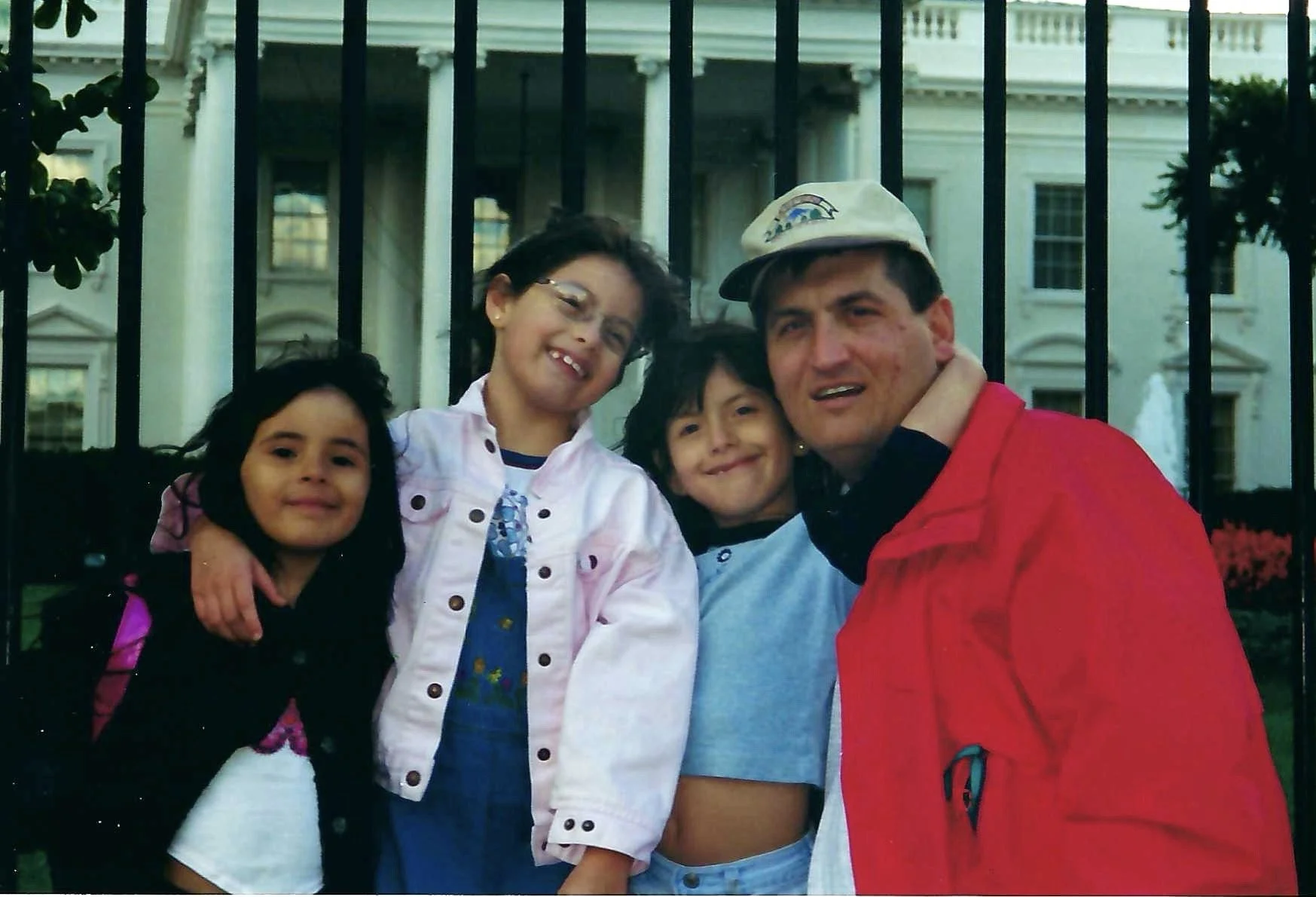 A man with a baseball cap and a red jacket is kneeling in front of a black fence with three little girls, all smiling and hugging him, in front of the White House.