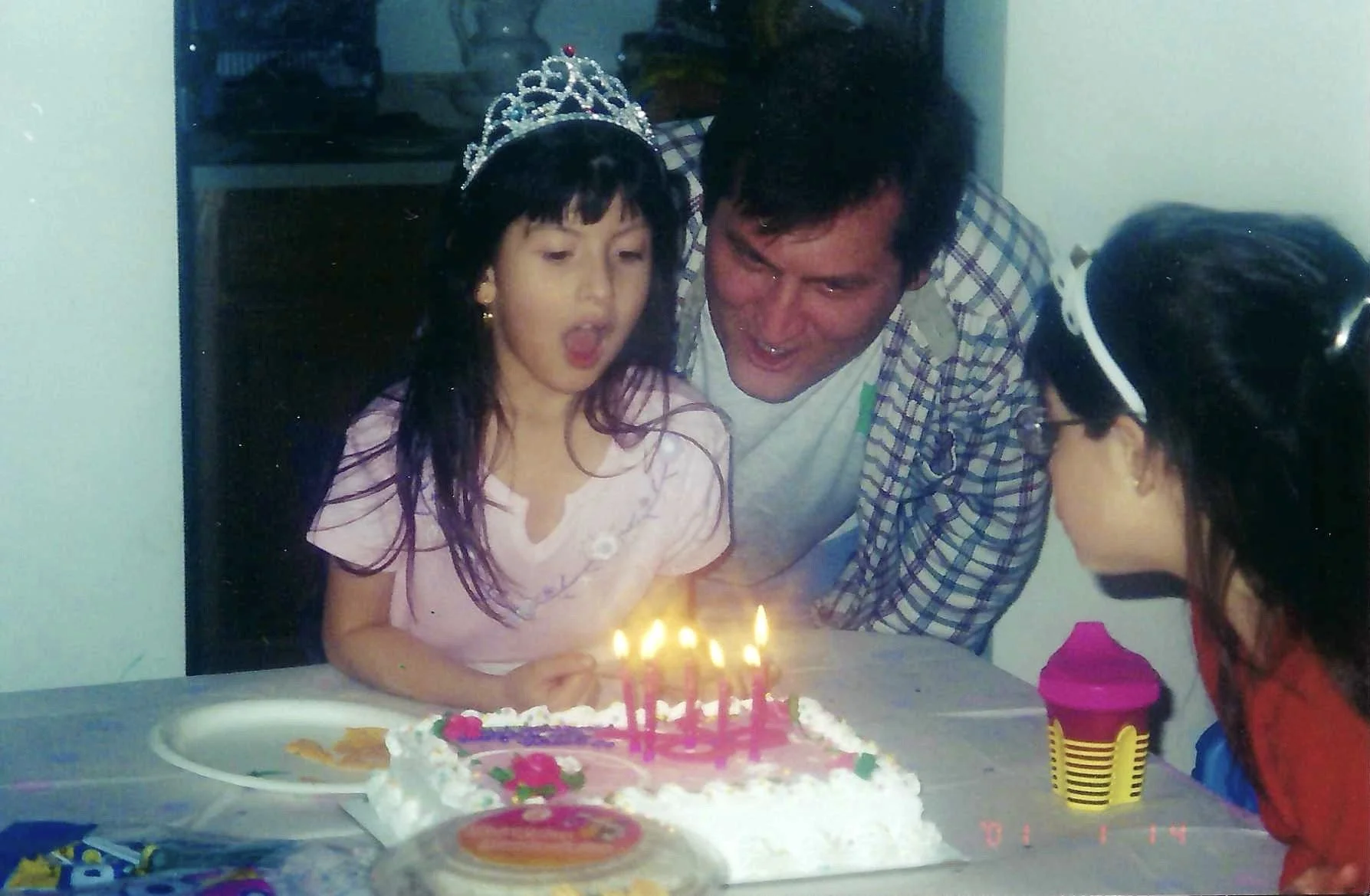 Young girl wearing a tiara and birthday girl, blowing out candles on her birthday cake surrounded by family members in a home setting.