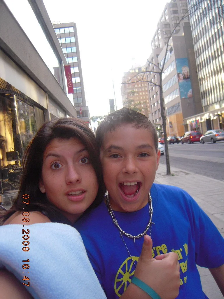 A woman and a boy taking a selfie on a city sidewalk, smiling and making funny faces. The woman has dark hair, and the boy has short hair, wearing a blue t-shirt and a beaded necklace. Buildings and cars are visible in the background.