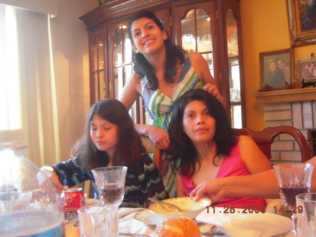 Three women sitting at a dining table with food and drinks, one young girl sitting with her eyes closed, and a woman standing behind them, smiling, in a warmly lit room with a wooden china cabinet, family photos, and a brick fireplace in the backgrou