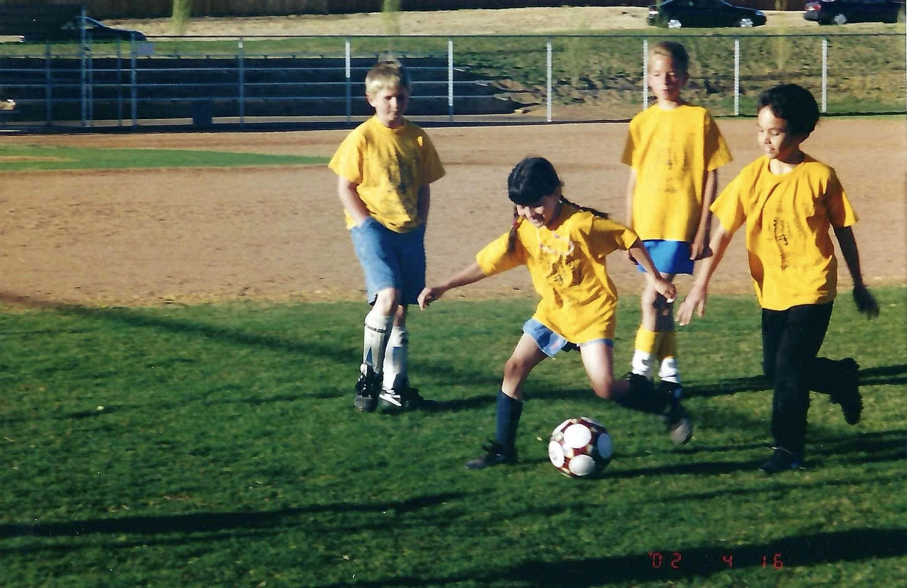 A group of children in yellow shirts playing soccer on a grassy field, with some children watching and a fence in the background.