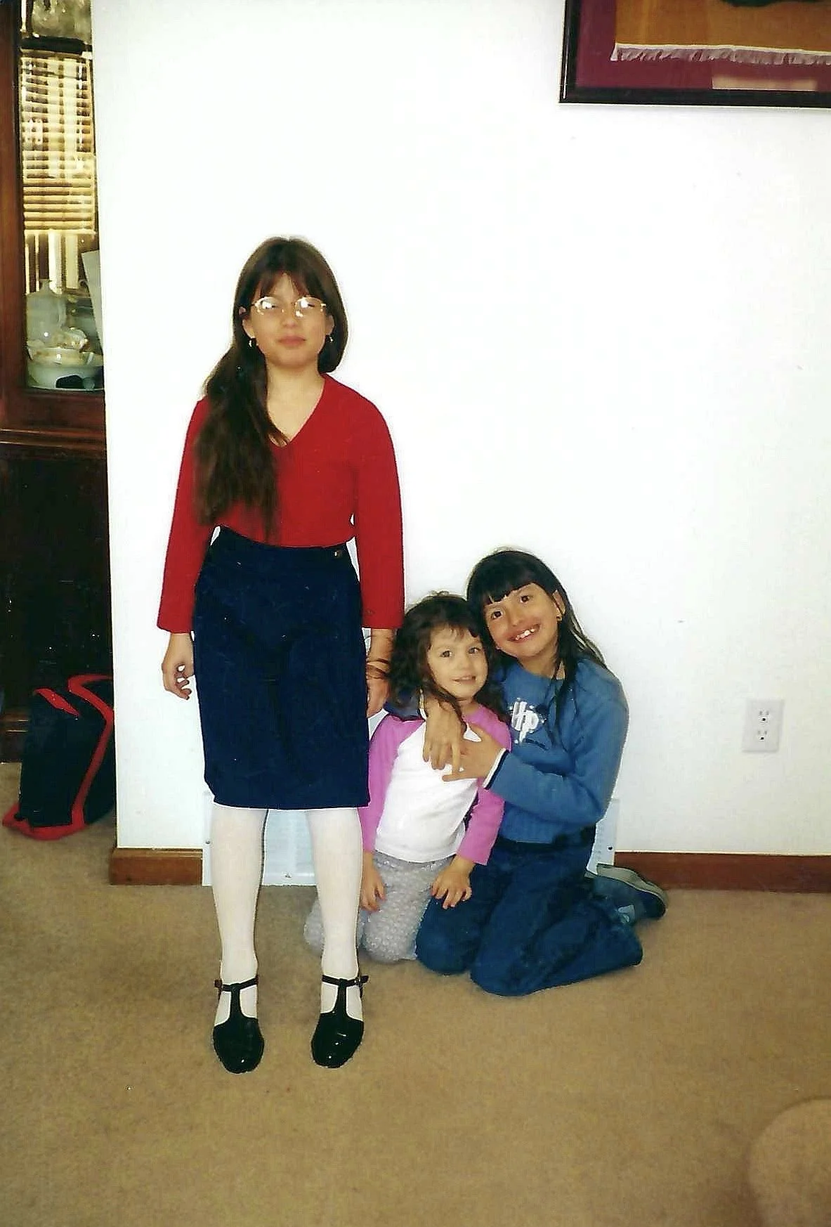 A woman and three young girls posing in front of a white wall, with a framed picture hanging above. The woman is standing while the girls are kneeling or sitting on the floor, smiling and hugging each other.