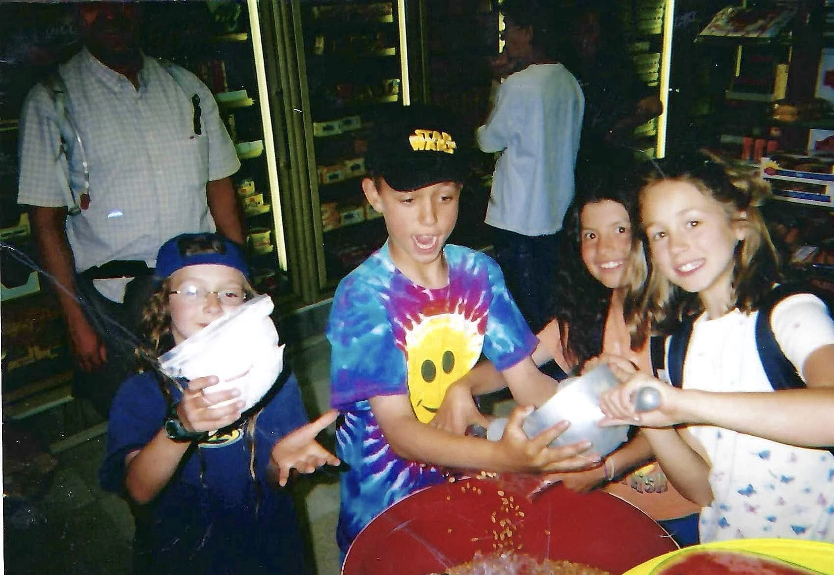 Four children with excited expressions gather around a table with food, while two adults stand behind them in a snack aisle.