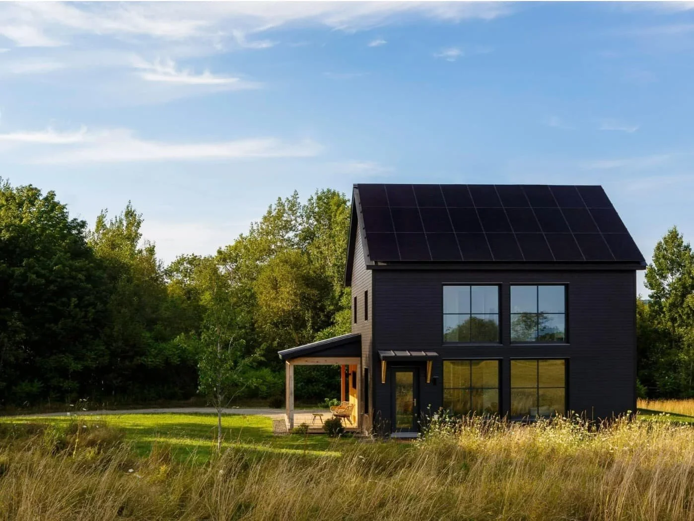 Black two-story house with solar panels on roof, surrounding green trees, and a grassy yard under a partly cloudy sky.