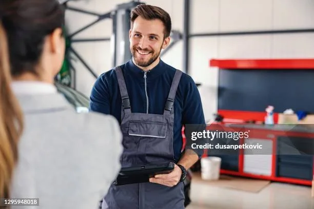 A smiling man in a mechanic's uniform holding a clipboard, talking to a woman in a workshop.