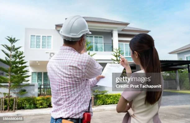 Two people, one wearing a hard hat and holding a clipboard, and the other pointing, standing in front of a modern two-story house with large windows and a balcony.