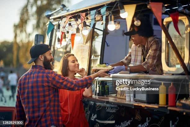 A man and a woman ordering food from a food truck decorated with string lights and colorful bunting, outdoors during daytime.