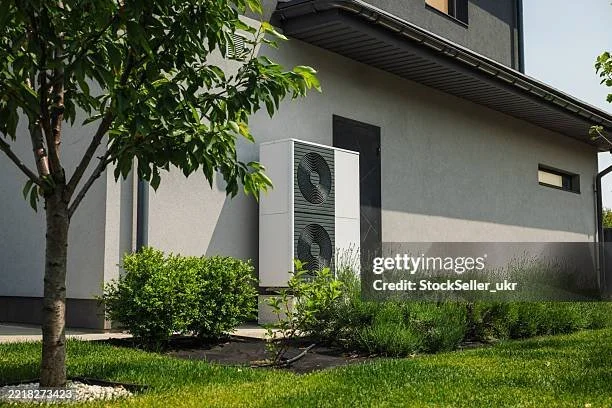A modern air conditioning unit installed on the exterior of a gray house, surrounded by green shrubs and grass, with a tree on the left side and a small window on the right.