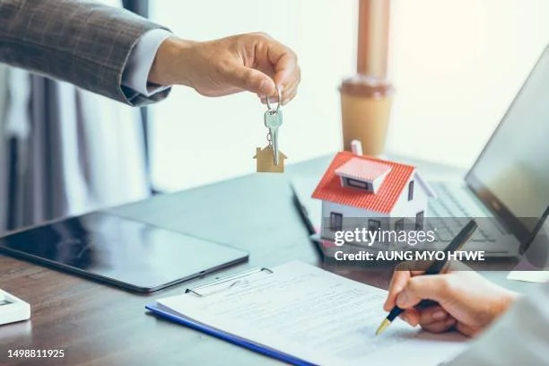 Person handing over a house key to another person in an office setting with a house model, laptop, tablet, and documents on the desk.