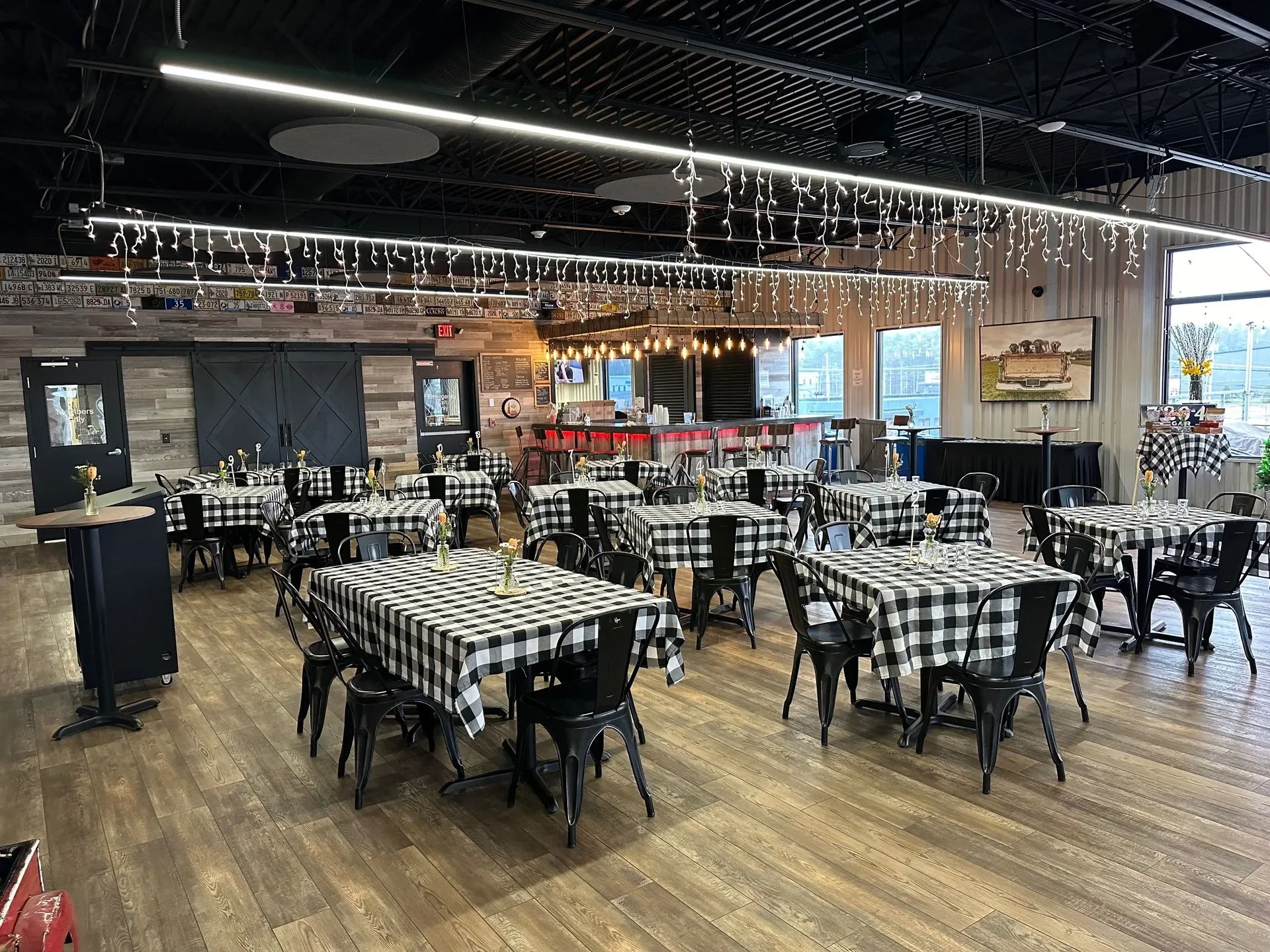 Interior of a restaurant with checkered tablecloths, black chairs, string lights, a bar area with barstools, and a wall with license plates and artwork. Large windows provide natural light.