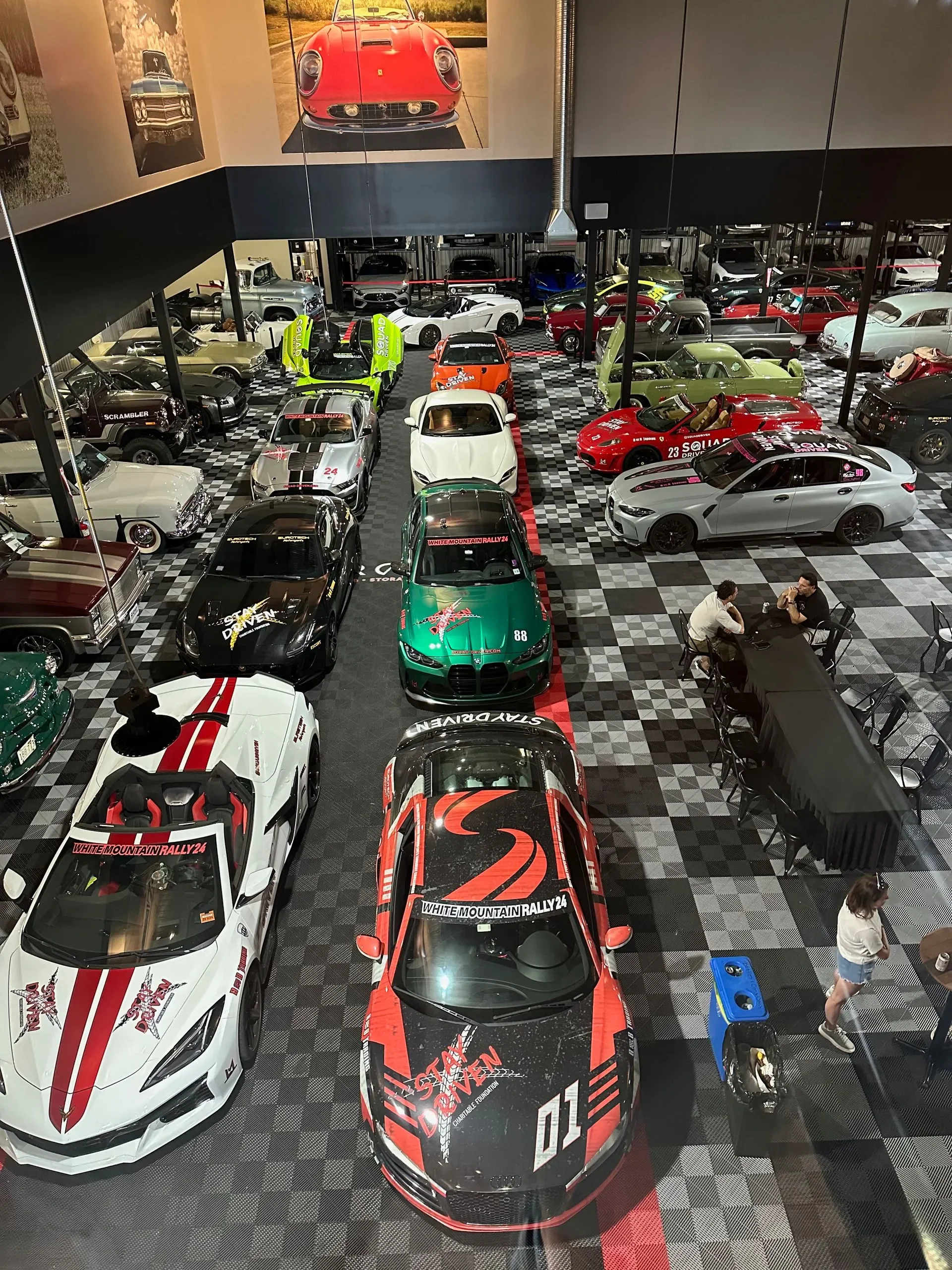 A large collection of cars on display in an indoor exhibition, with some cars decorated with rally-themed graphics and text, and a few people sitting at tables.