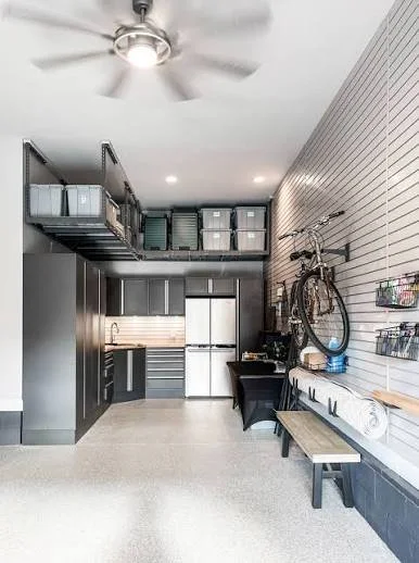 A well-organized garage with gray cabinetry, a refrigerator, and wall-mounted storage. There is a wooden bench, a bicycle mounted on a pegboard, and industrial-style shelving with storage bins.