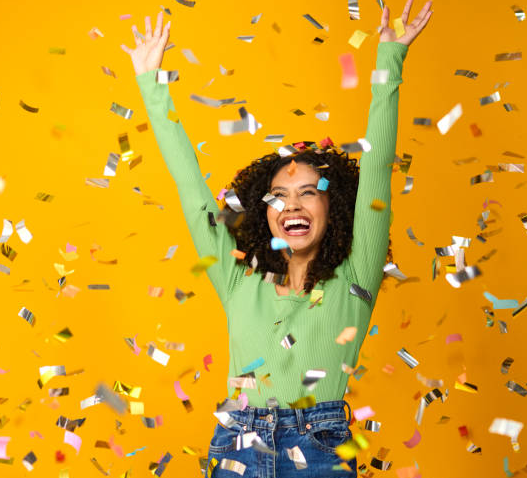 Woman celebrating with arms raised amid falling confetti against a yellow background.