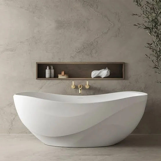 Modern, white freestanding bathtub with a curved design, mounted on a light-colored floor against a textured gray wall, with a wooden shelf above holding toiletries and decorative items.