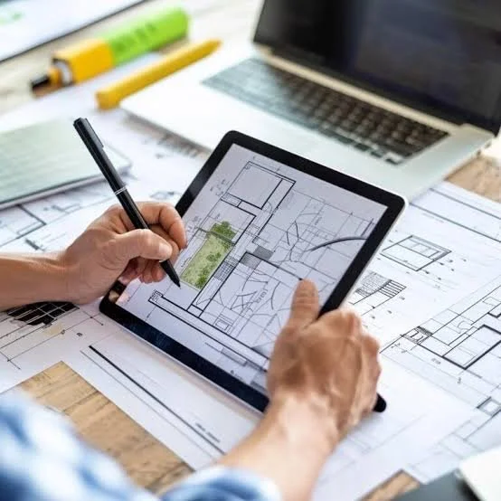 Person using a stylus on a tablet to view architectural designs, with blueprints, a laptop, and drawing tools on a desk.