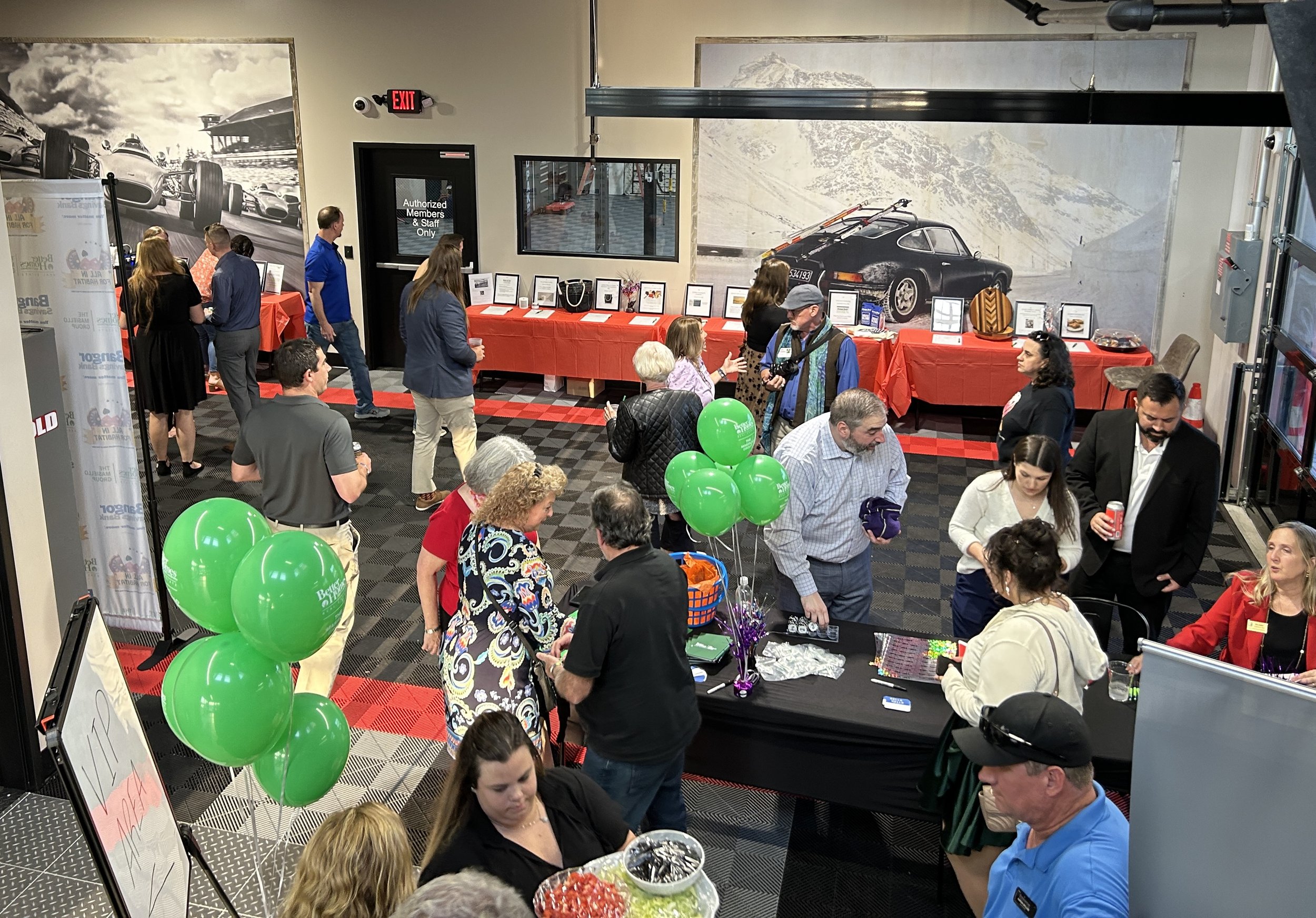 An indoor event with people gathered around tables that have framed photos and items on display. There is a large wall mural of a racing car and mountain landscape. Green balloons are in the foreground.