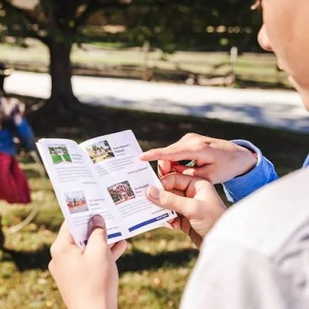 Person holding a folded brochure or map outdoors during daytime, with trees and a walking path in the background.