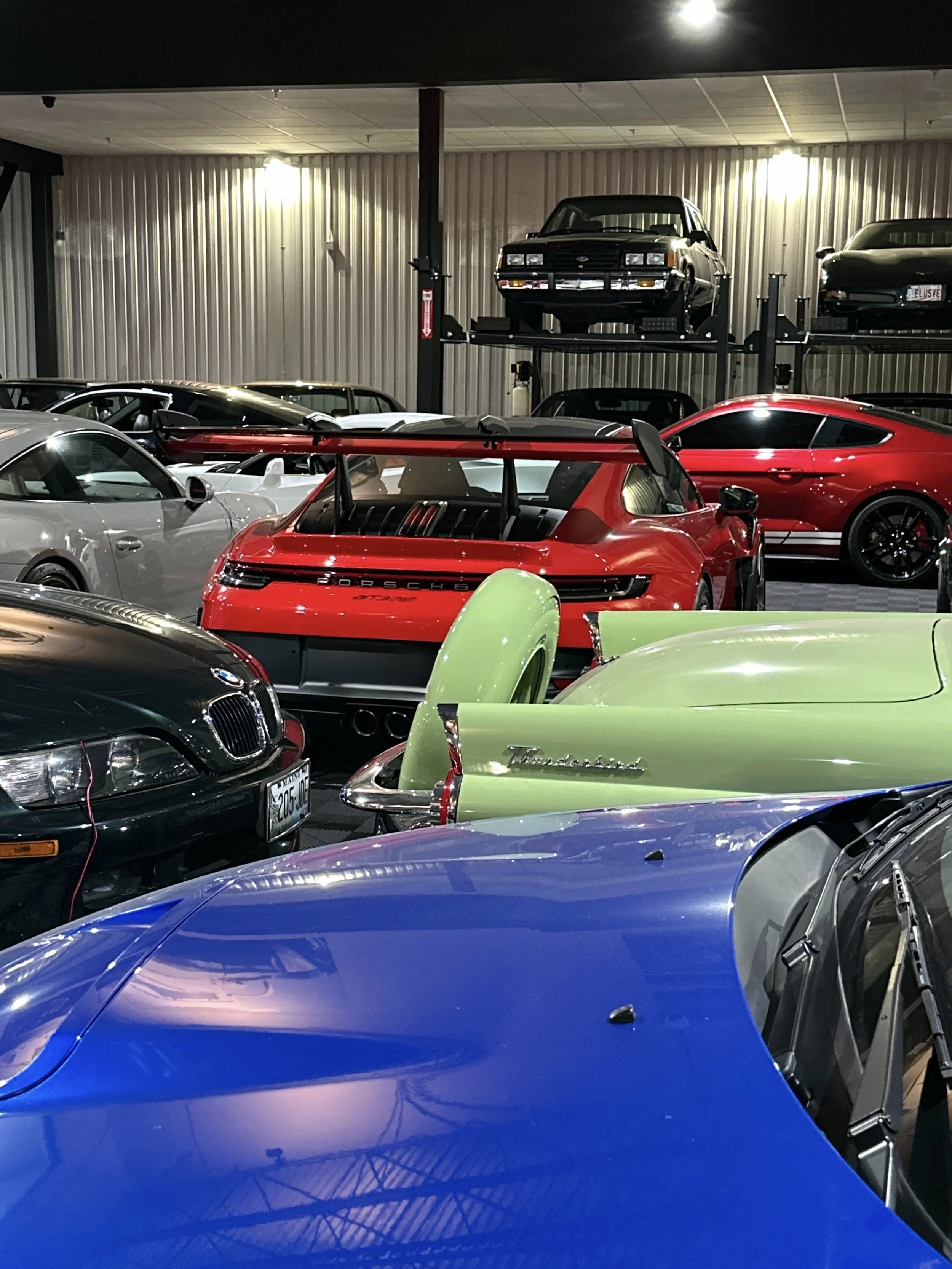 Indoor garage with classic cars, including a blue car in the foreground, a green car with a noticeable rear wing, a red Porsche 718, and a black BMW. Elevated cars are in the background on metal racks.