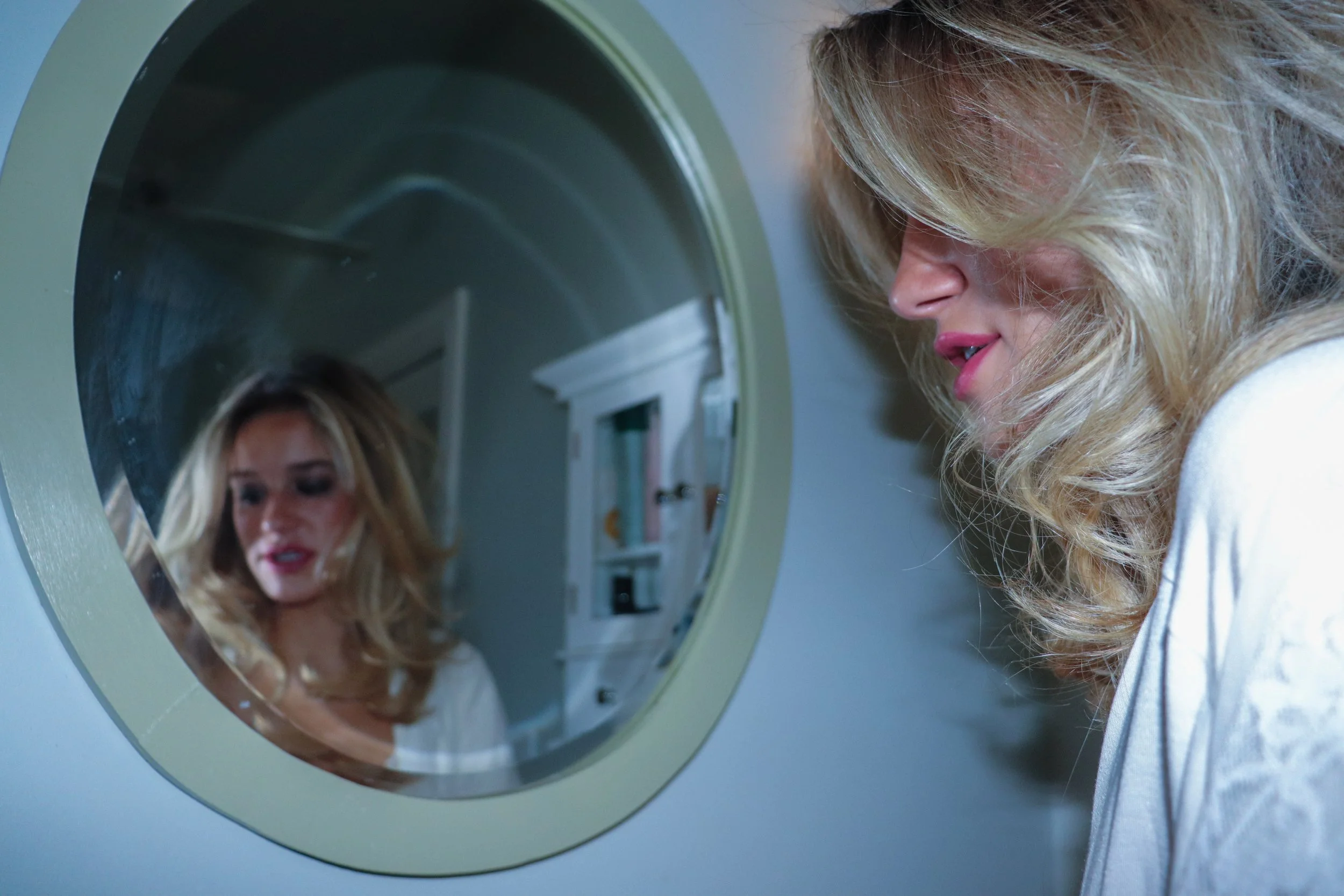 A blonde woman with smokey make up stands facing a mirror with her reflection visible and looking away.