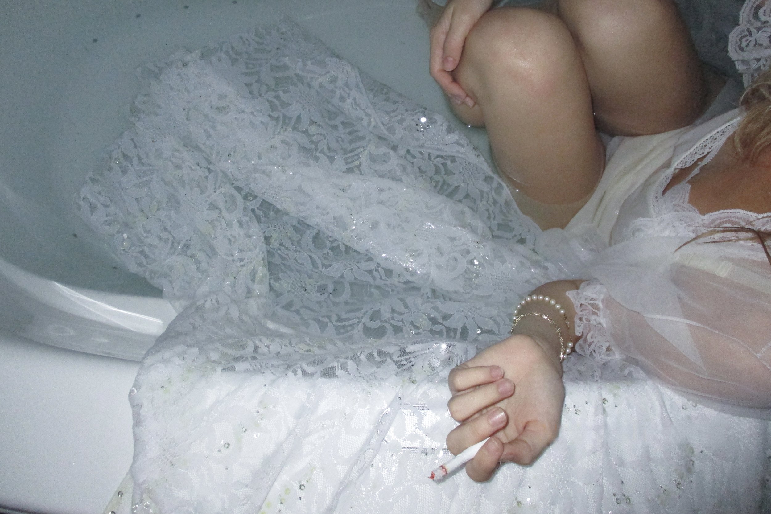 Birds-eye view of a woman draped in sheer and lace white garments, sitting in a half-filled bathtub, smoking a cigarette.