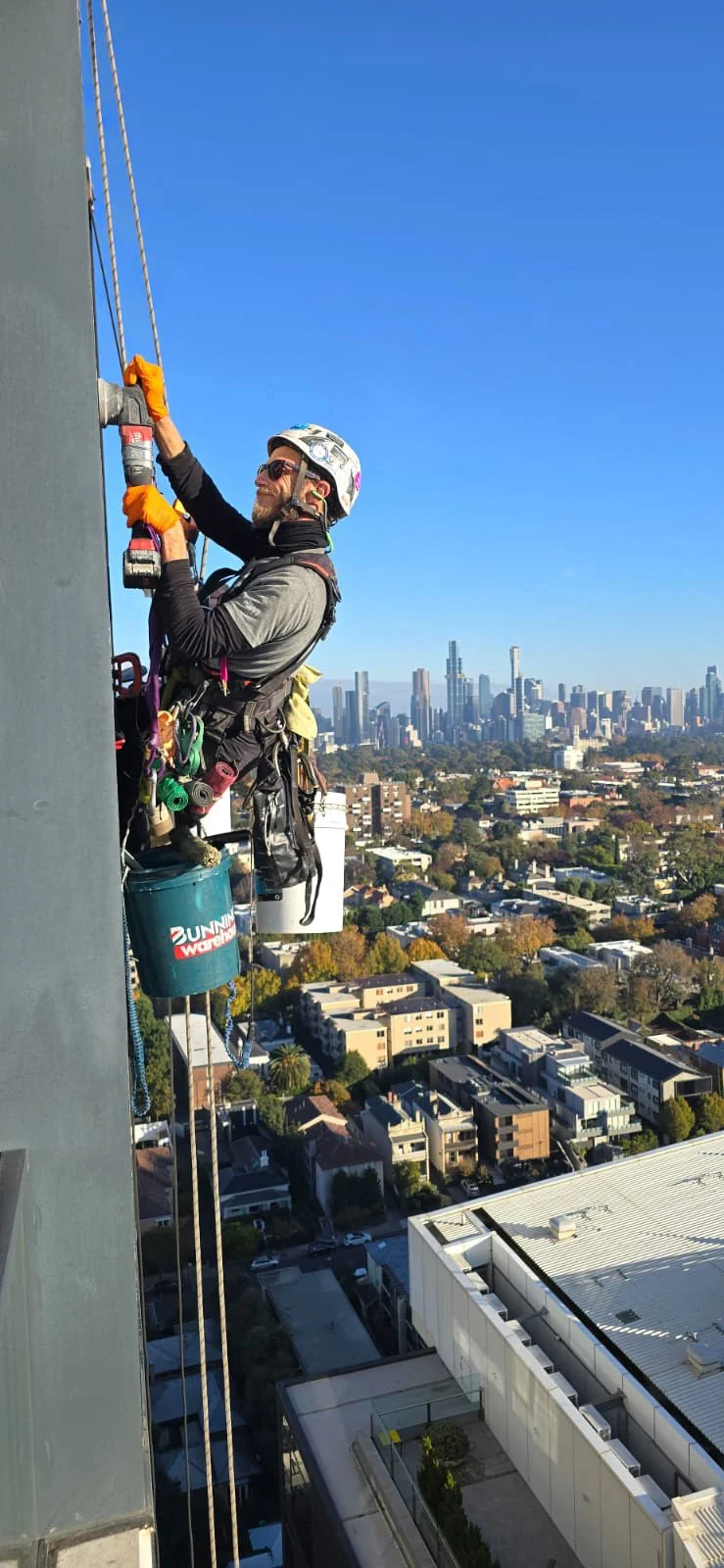 A high access rope technician, secured with safety harnesses, working on a building's exterior with a city skyline in the background.