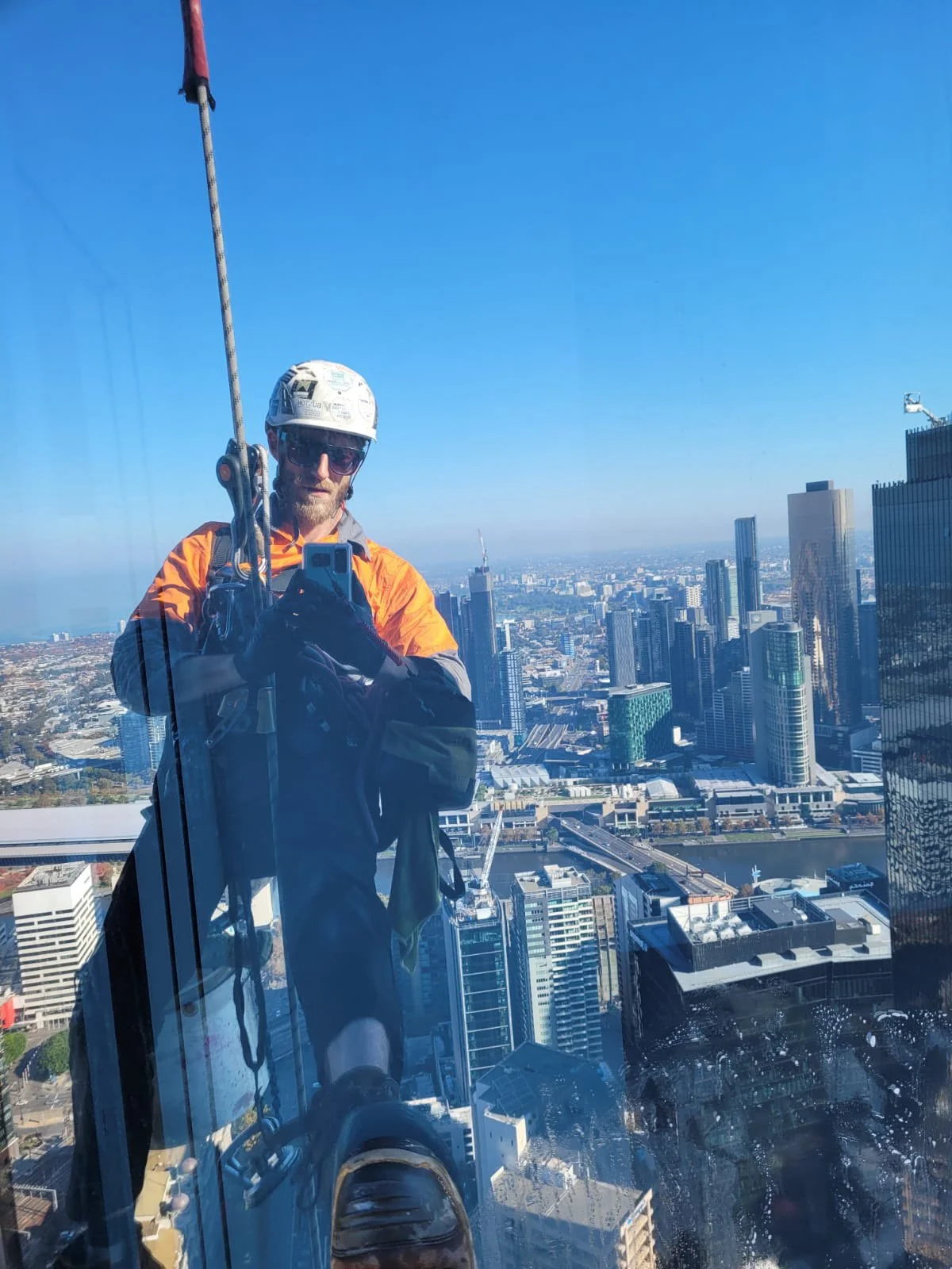 A high access rope technician, in safety gear, helmet, sunglasses, and orange hi-vis, suspended on ropes with a tall city skyline and river in the background, reflected in a glass panel.