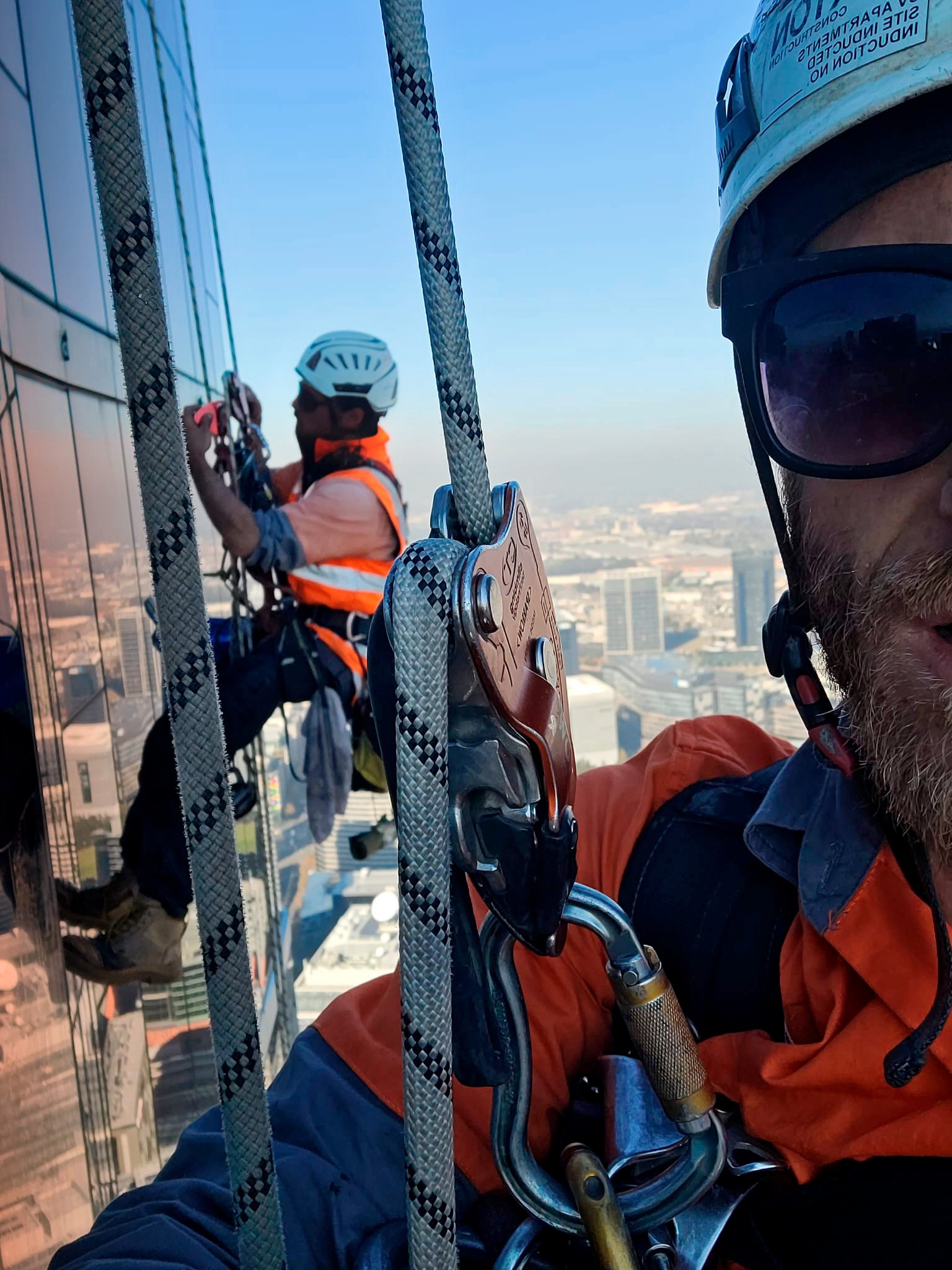 A high access rope technician, in an orange and blue jacket with a helmet and harness, working on a high-rise building with another worker in the background adjusting safety equipment, with city skyline and tall buildings visible in the distance.