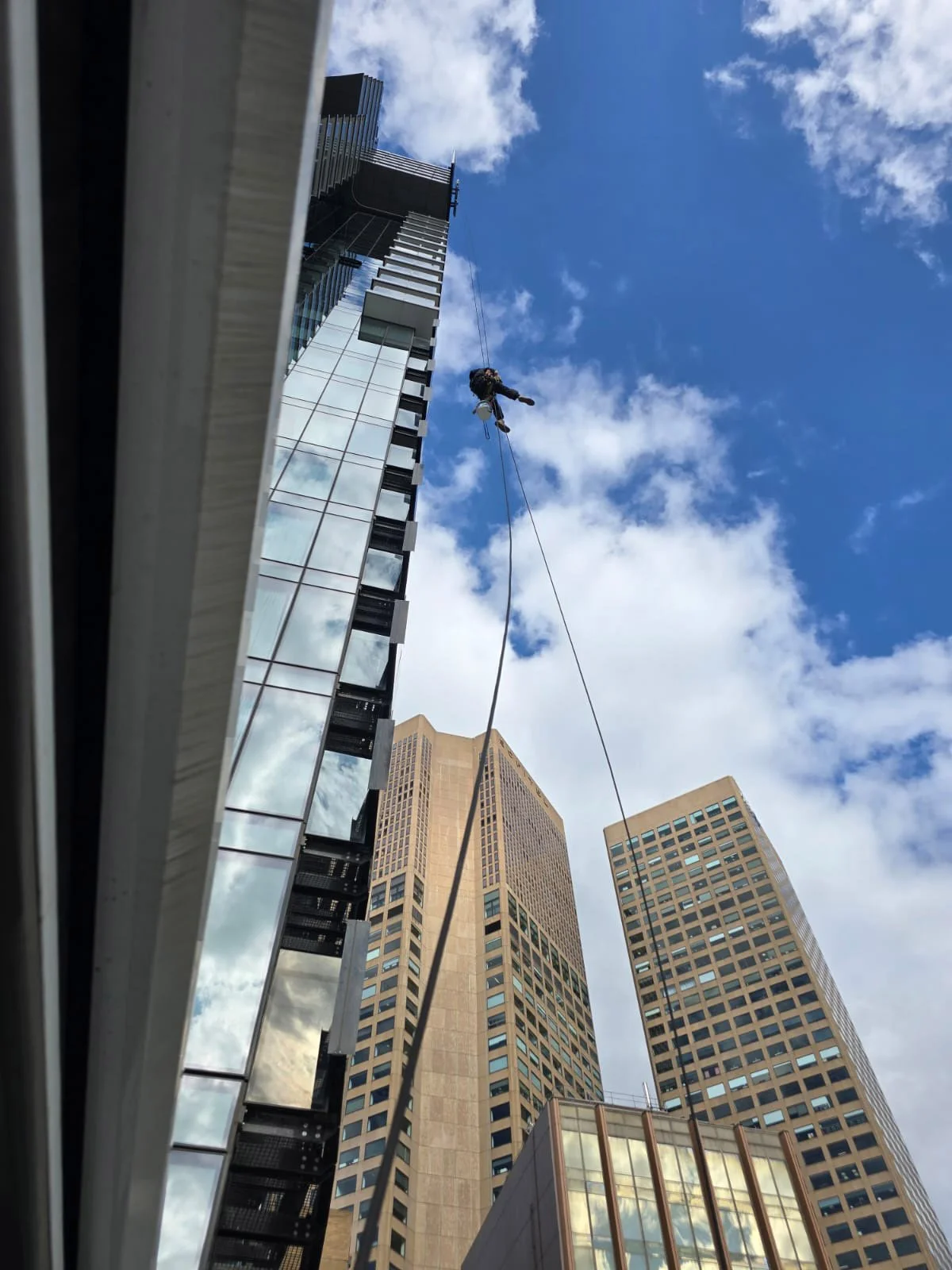 A worker is suspended by a harness and rope, working on the exterior of a high-rise building in a cityscape with other tall buildings under a partly cloudy sky.
