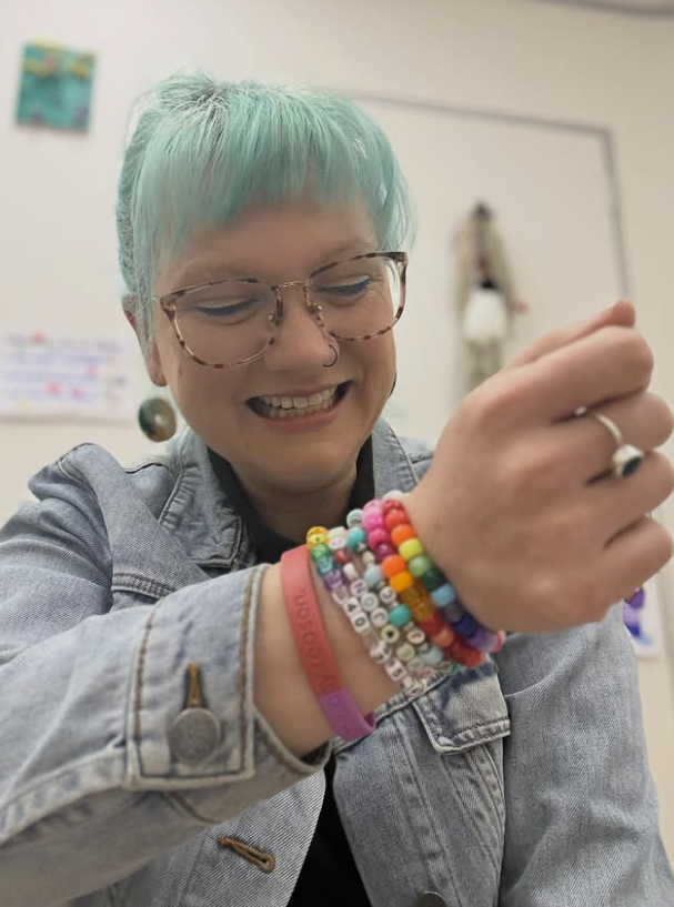 Bri smiling while looking down, wearing glasses, a denim jacket and a colourful collection of beaded bracelets.