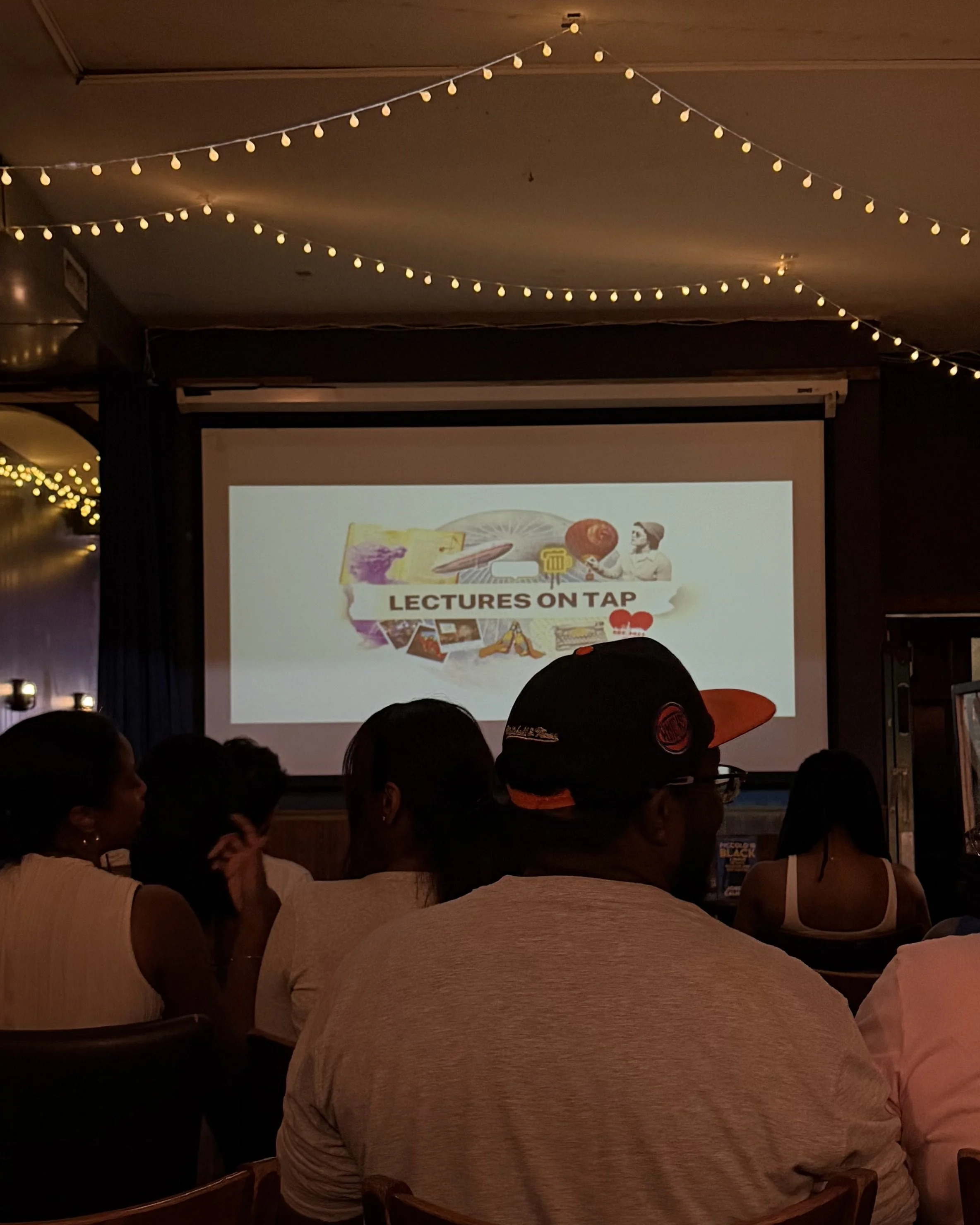 View of a dark room with a large projector screen displaying a presentation titled 'Lectures on Tap' with various images. The room has string lights hanging from the ceiling, and people are seated watching the presentation.