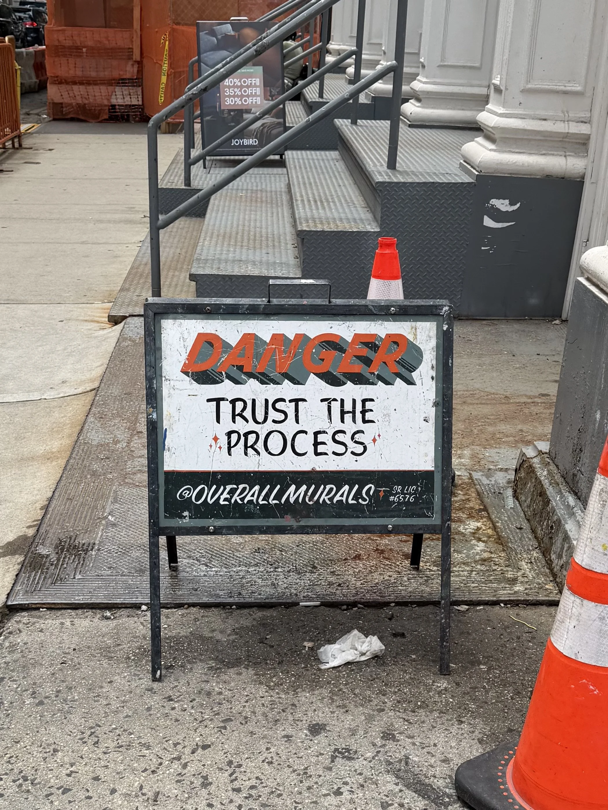 Street sidewalk with safety cone and a sign that says "DANGER TRUST THE PROCESS @OVERALLMURALS".