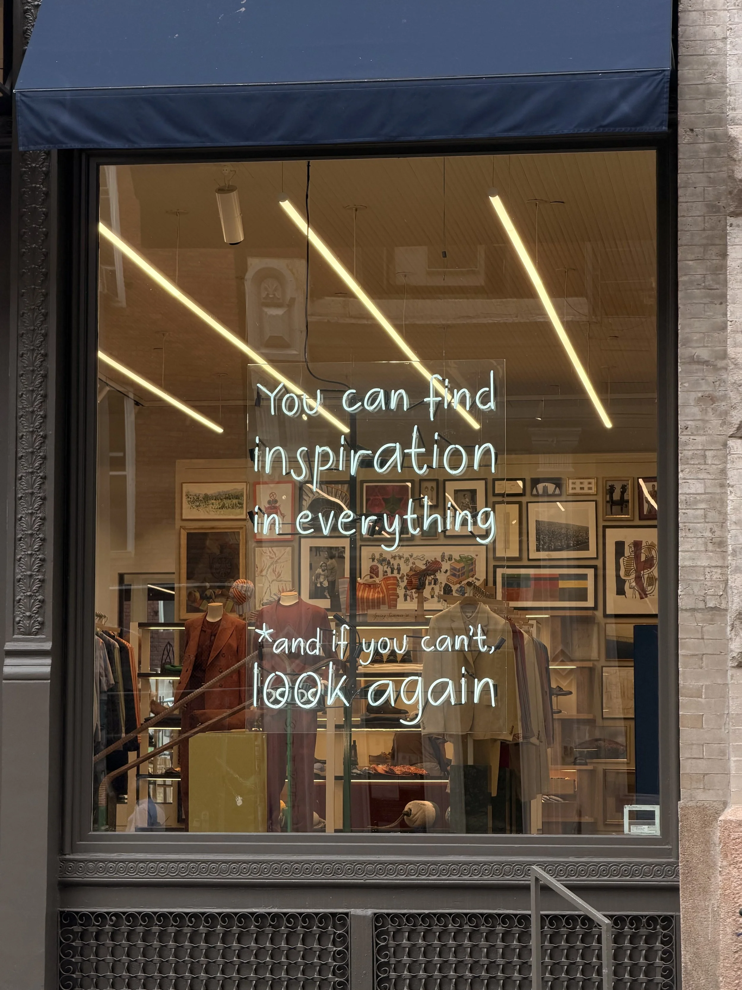 Store window with a neon sign that reads, "You can find inspiration in everything," and a smaller note below that says, "and if you can't, look again." The window displays clothing mannequins and framed artwork inside.