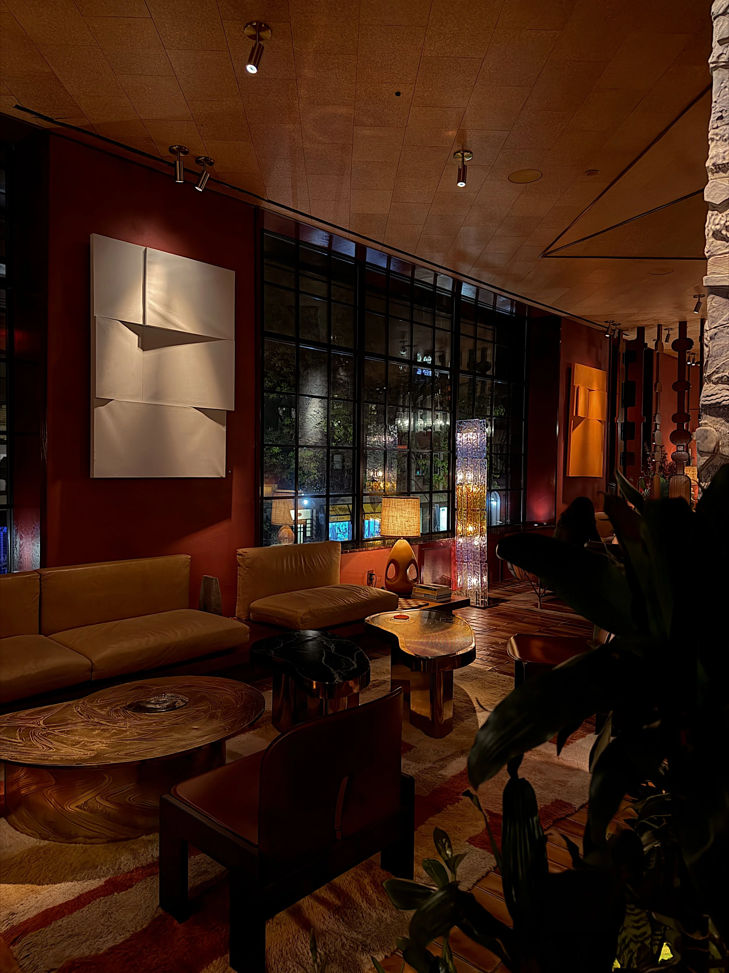 Interior of a cozy, stylish living room with a beige couch, wooden coffee tables, decorative lighting, large window with black grid, and red and wooden decor accents.