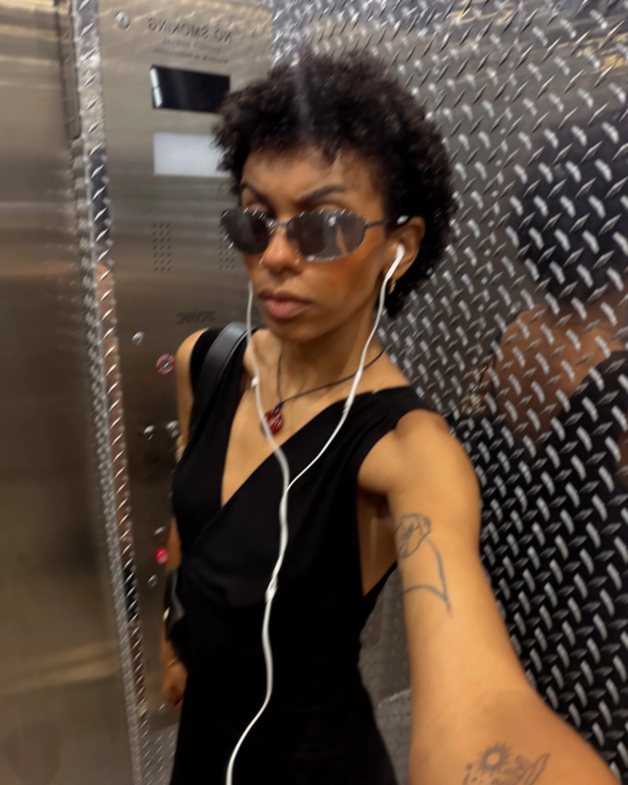 A woman with short curly hair, wearing sunglasses, black tank top, and earphones, standing in an elevator with metal diamond plate walls.
