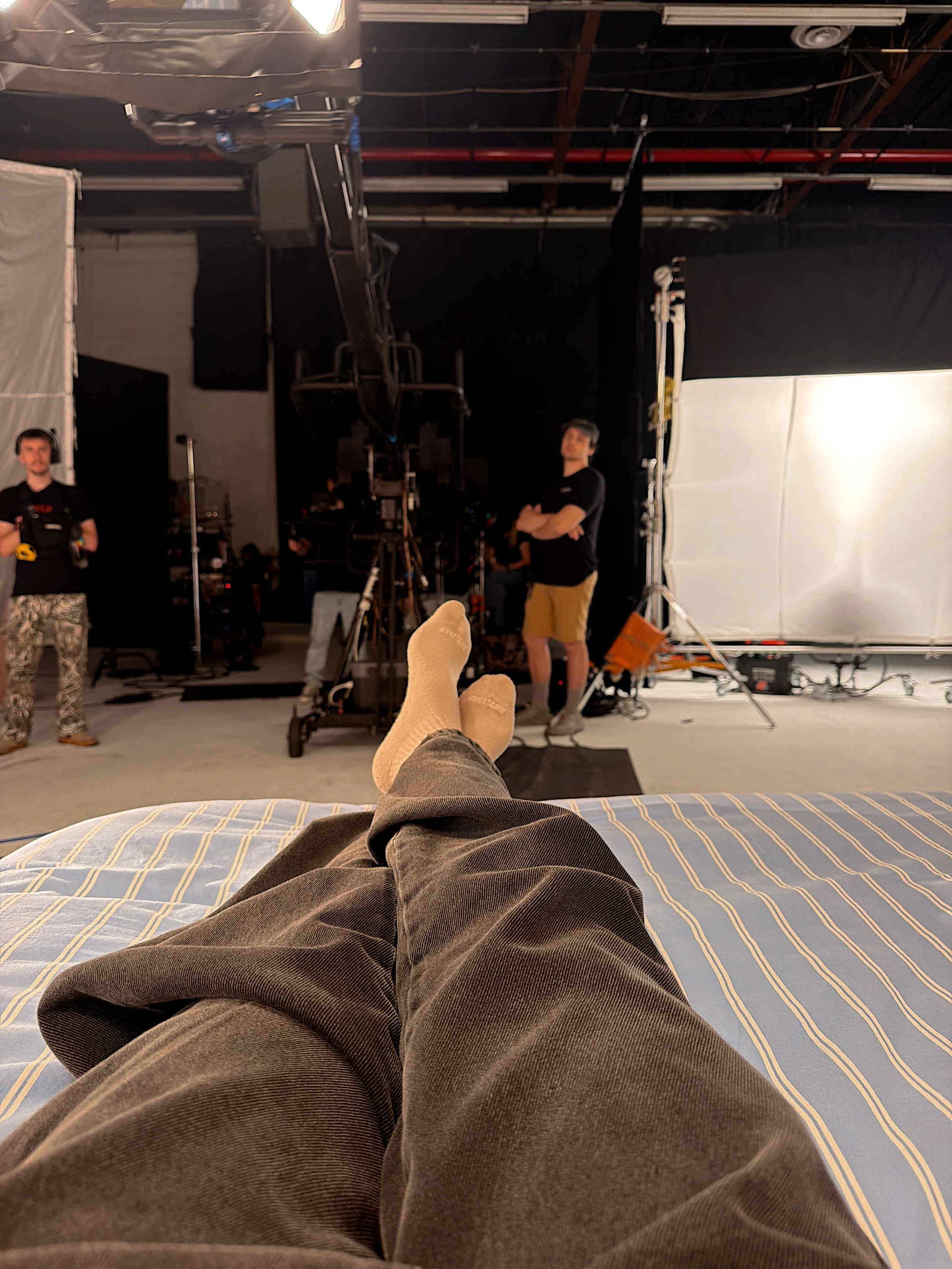 Person lying on a bed with feet and legs visible in the foreground, facing a film or photo shoot setup with crew members, lighting, and equipment.