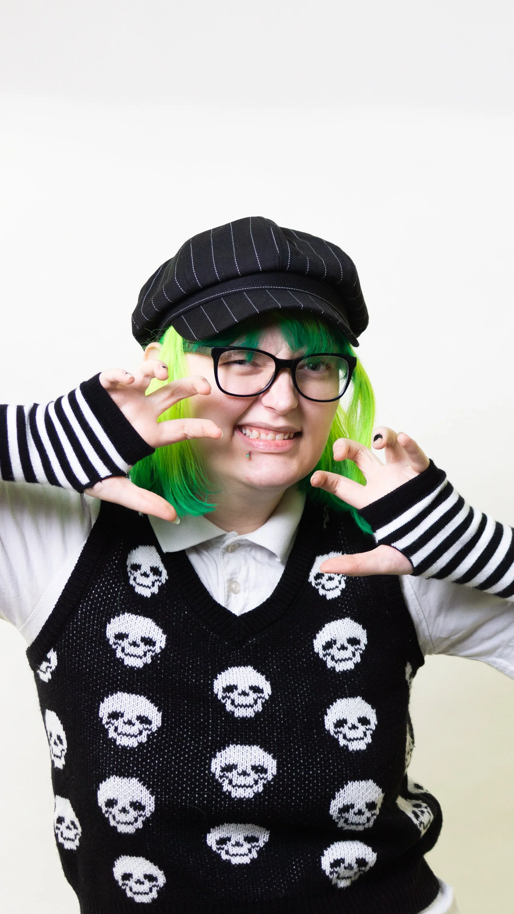 Photograph of Lilly Day, she has green hair, black glasses, a black hat with white pinstripes, a white collared shirt, and a black vest with white skulls, smiling and making a claws gesture with fingers.