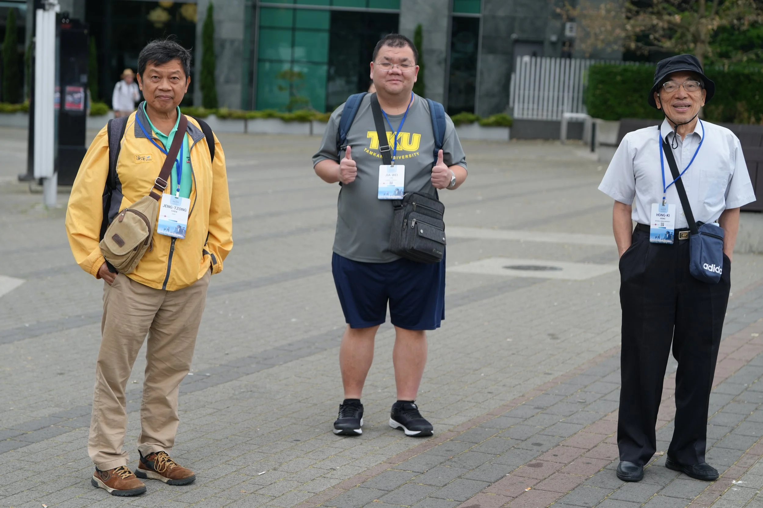 Three men standing outdoors in front of a modern building, wearing casual clothes and backpacks, with name tags hanging around their necks.