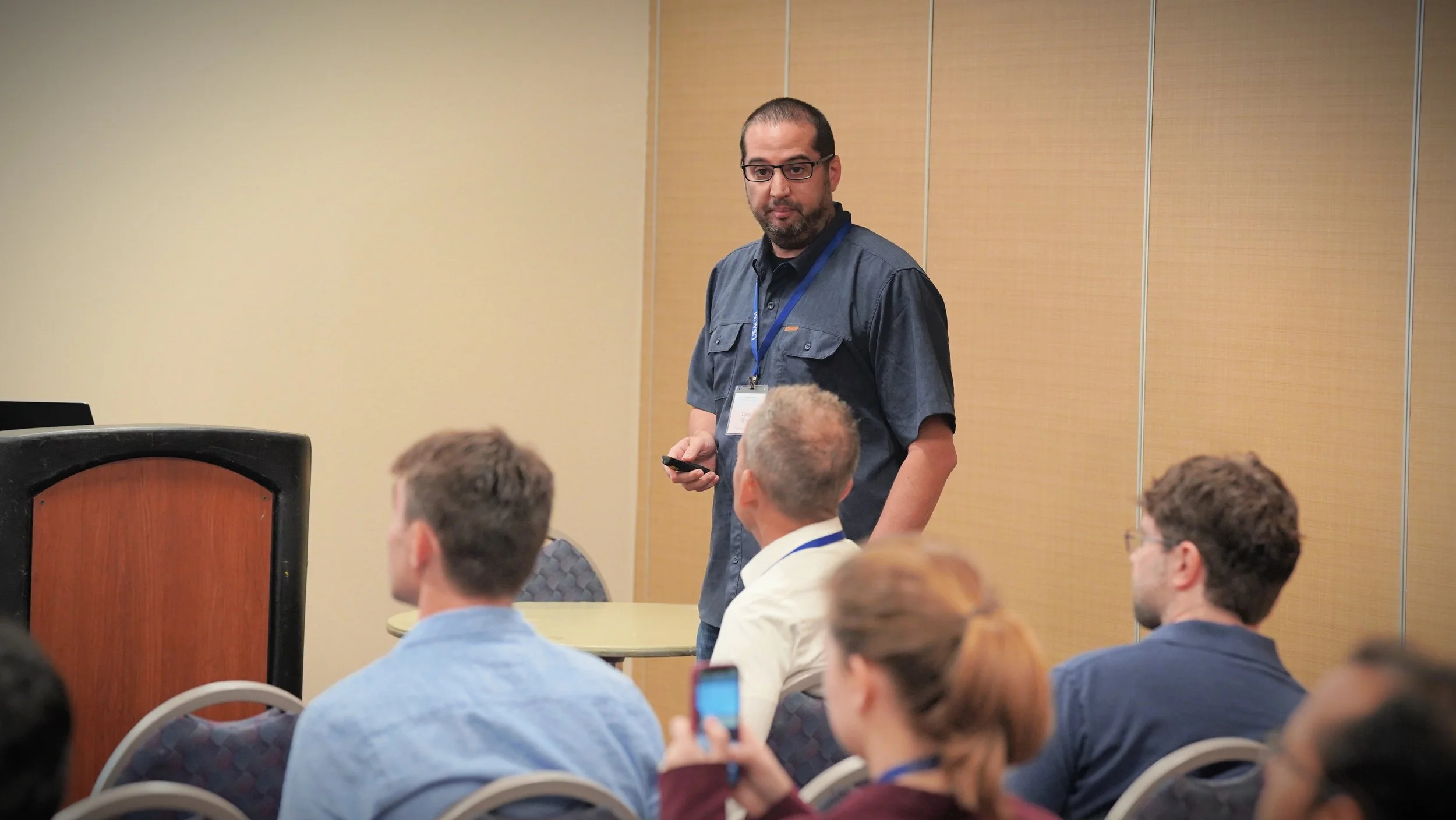 A man with glasses and a beard, wearing a dark shirt and conference badge, standing in a conference room and holding a phone. Several people are seated, some taking photos or listening.
