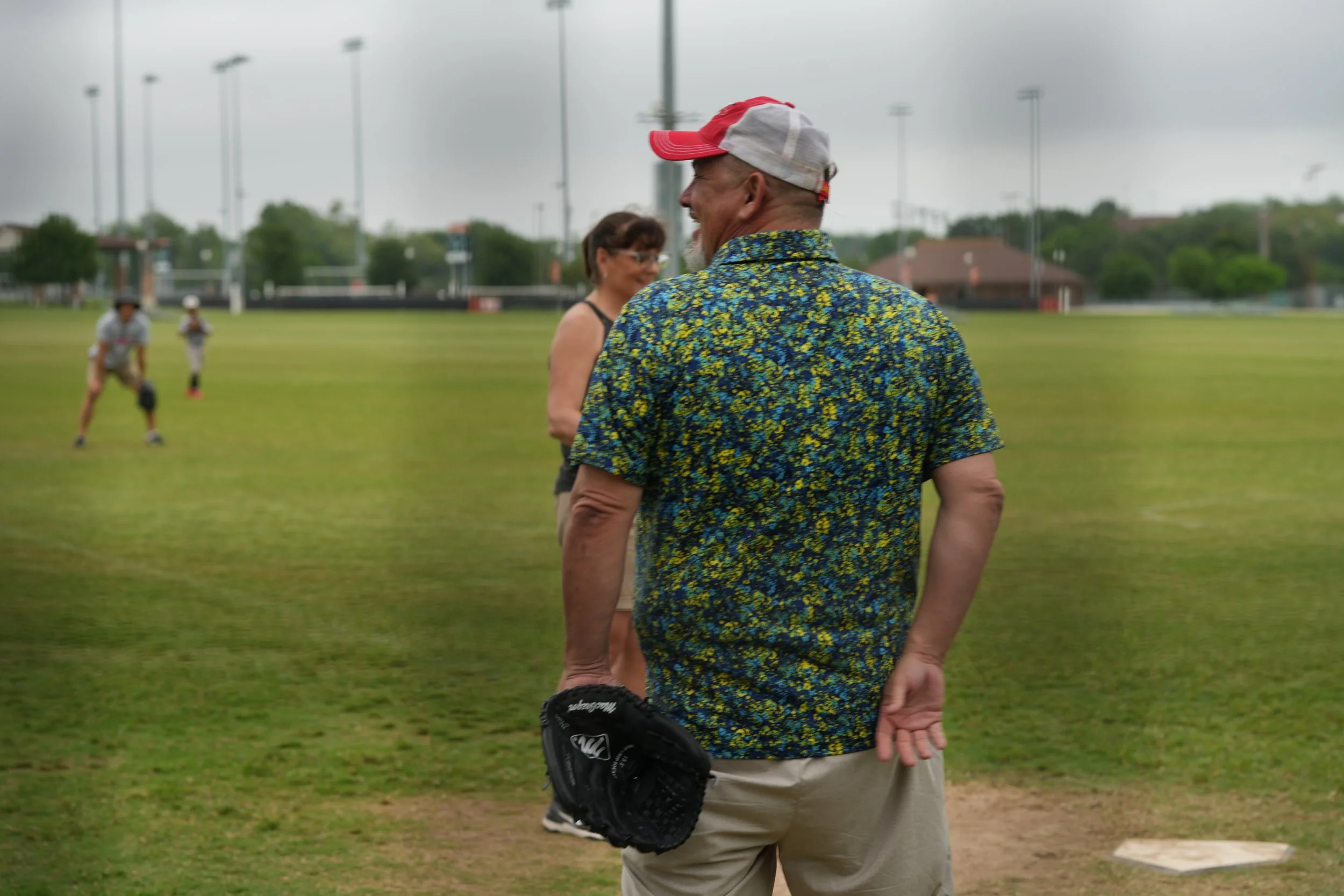 An older man in a colorful floral shirt and beige shorts, holding a baseball glove, stands on a grassy field near a baseball field. Behind him, a woman and children can be seen, with the children playing in the background. The scene appears to be overcast.
