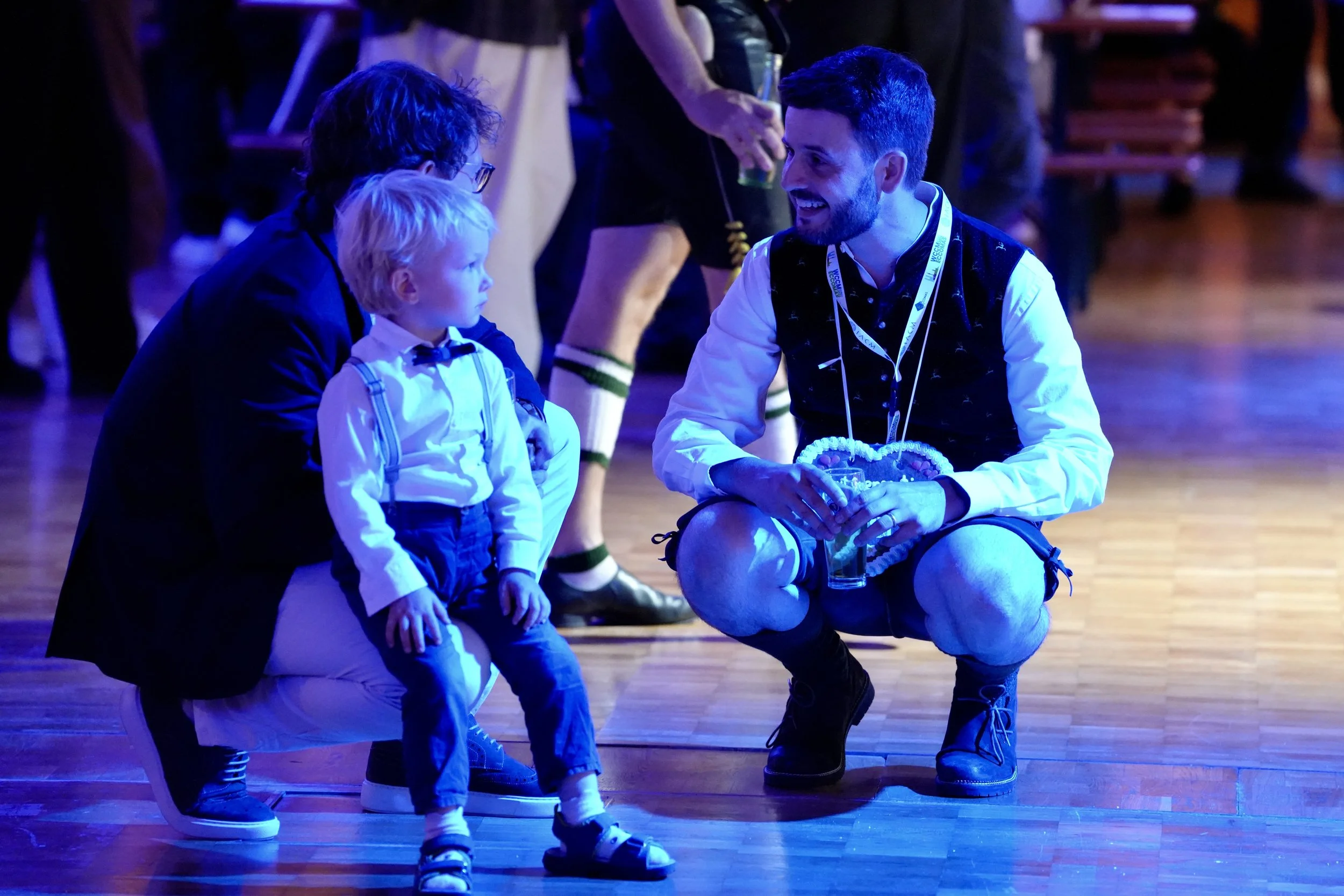 A man with a beard wearing a black vest and white shirt is kneeling and smiling while talking to two young boys sitting on a round object. One boy with glasses and the other with blond hair. The background is dark with an audience and performers, some in costume, illuminated by stage lighting.