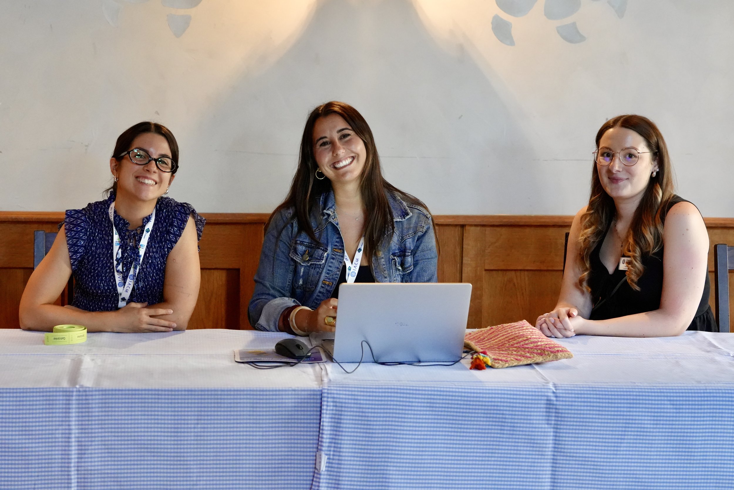 Three women sitting at a table in a well-lit room, smiling at the camera. They have notebooks, a laptop, and personal items on the table.