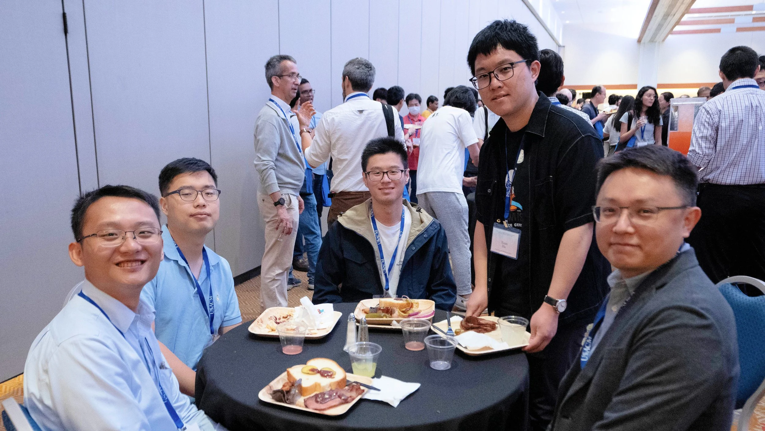 A group of people gathered around a table at a conference or event, with some eating and others standing and socializing in the background.
