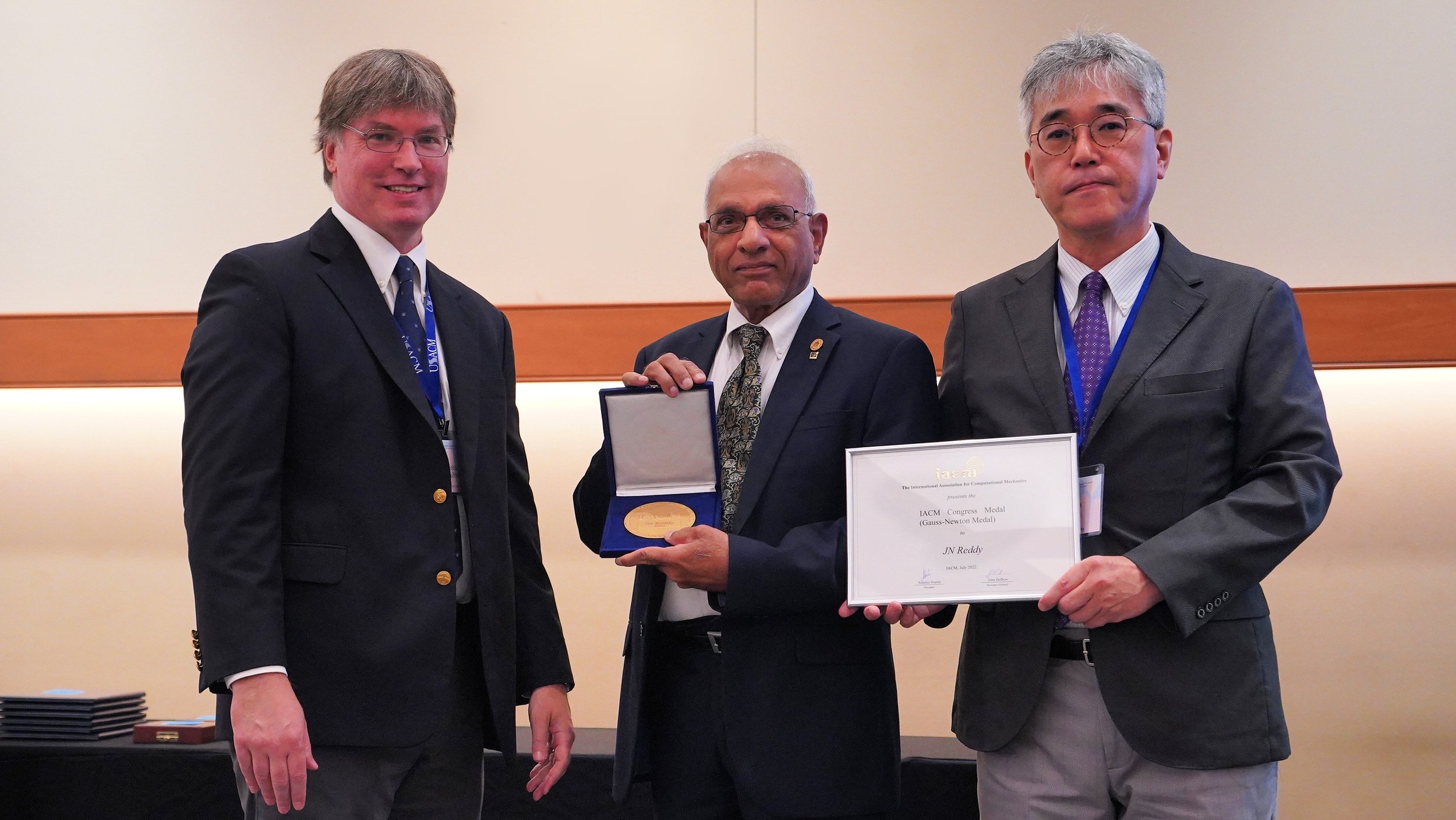 Three men in business suits at an award ceremony, one holding a certificate and the other holding a medal case, standing in front of a beige wall.