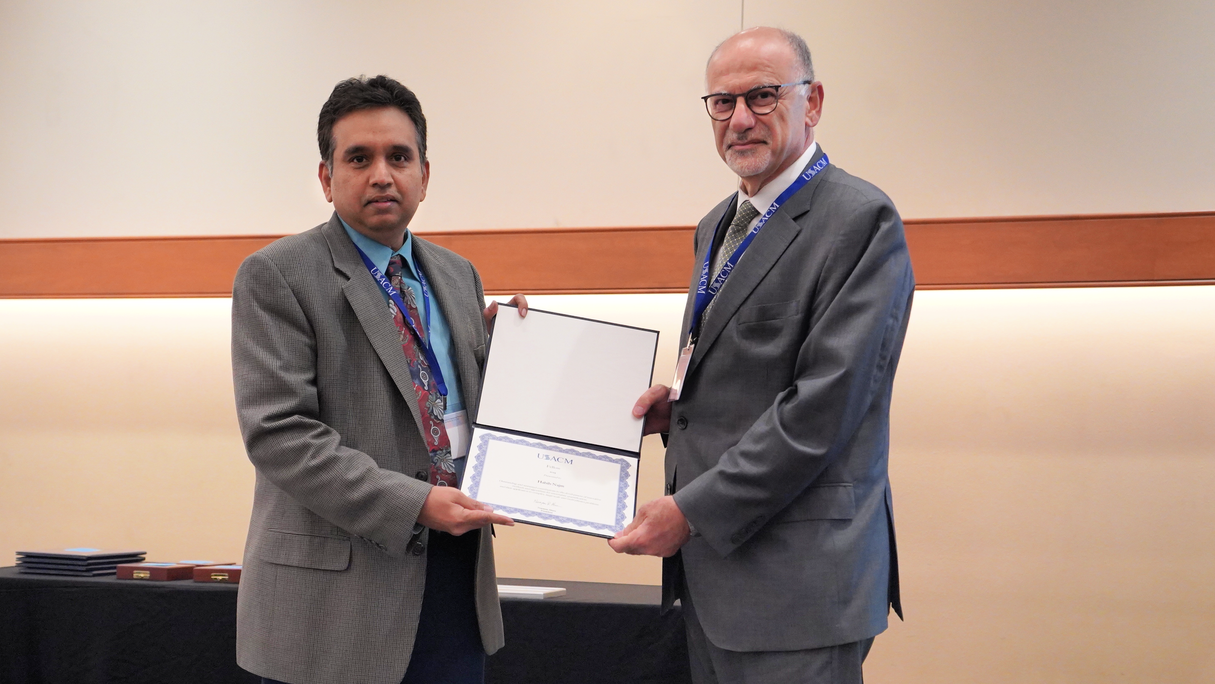 Two men in suits shaking hands and exchanging a certificate at an awards ceremony. One man is younger, with dark hair, wearing a gray checked suit and a colorful tie. The other man is older, with a beard, glasses, and a gray suit. Both are wearing lanyards with conference badges.