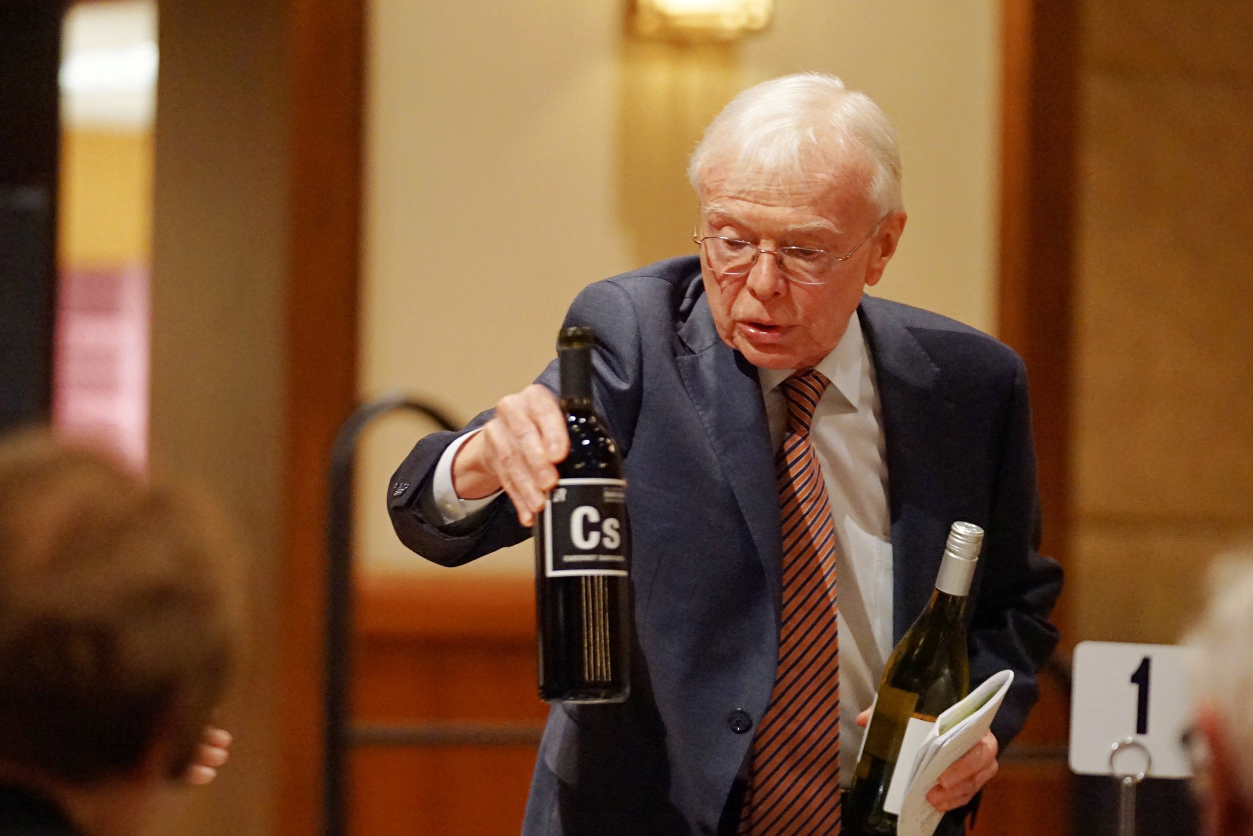 An elderly man in a suit and striped tie is pouring wine from a bottle labeled 'CS' at a table with wine bottles and documents, in a room with warm decor.