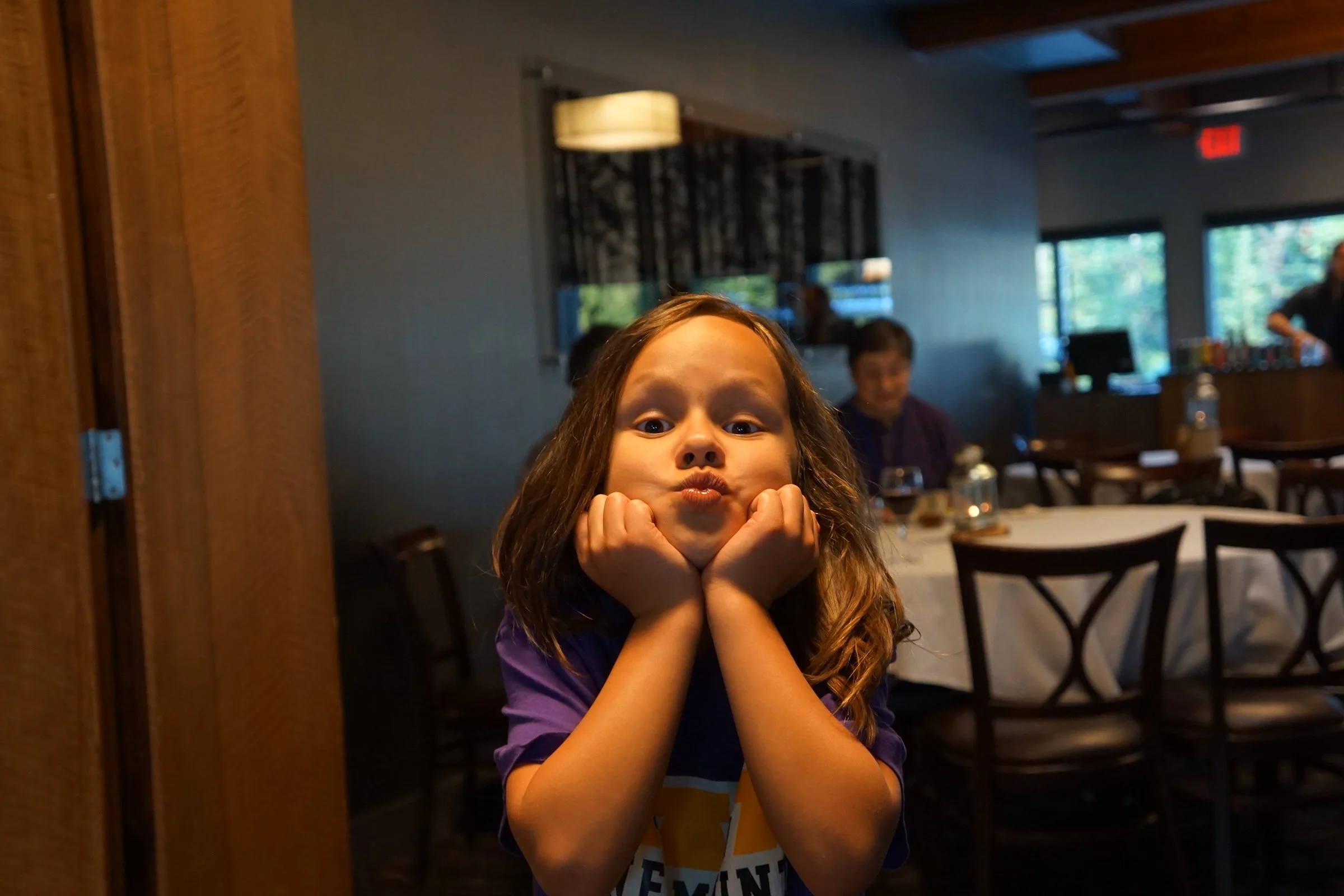 A young girl with light brown curly hair making a playful face with puffed cheeks and wide eyes, resting her chin on her hands in a restaurant or cafe setting.